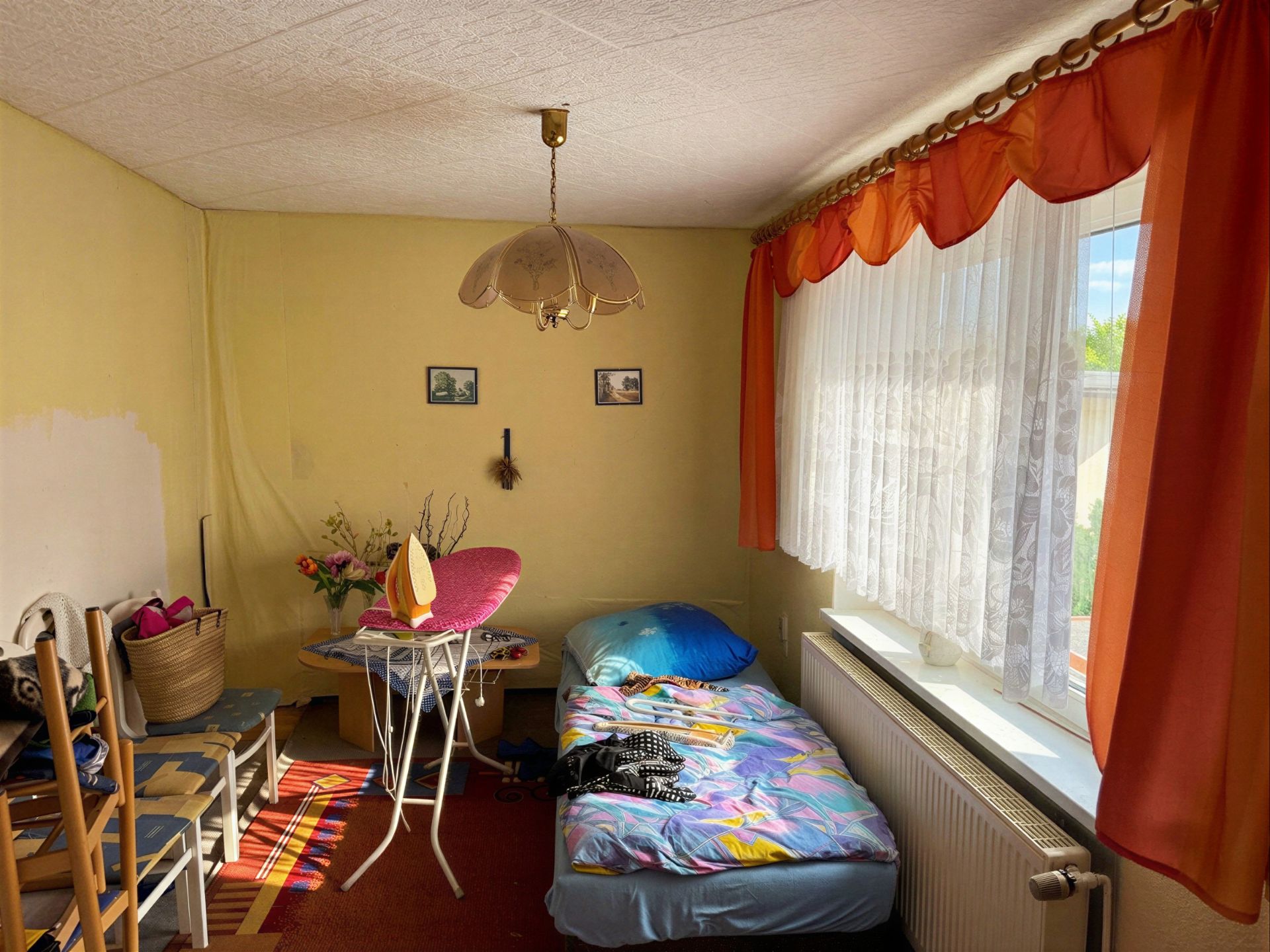 Kleines Zimmer mit Einzelbett, Stühlen, Tisch und großem Fenster mit orangenen Vorhängen.