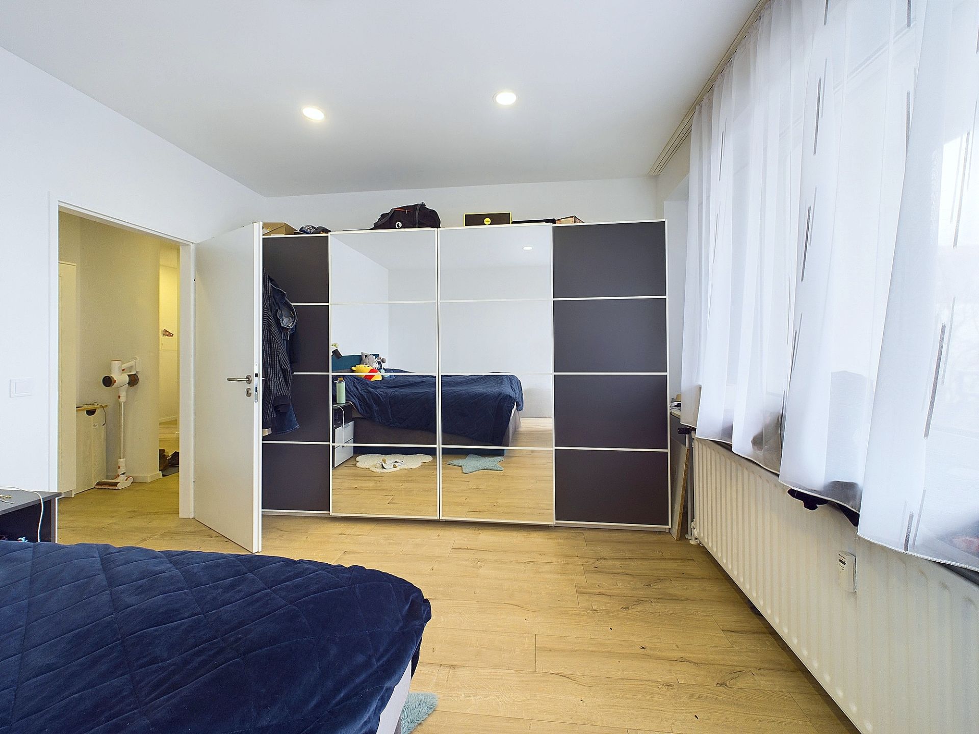 Schlafzimmer mit Kleiderschrank mit Spiegeln, Holzfußboden und großen Fenstern mit Vorhängen.