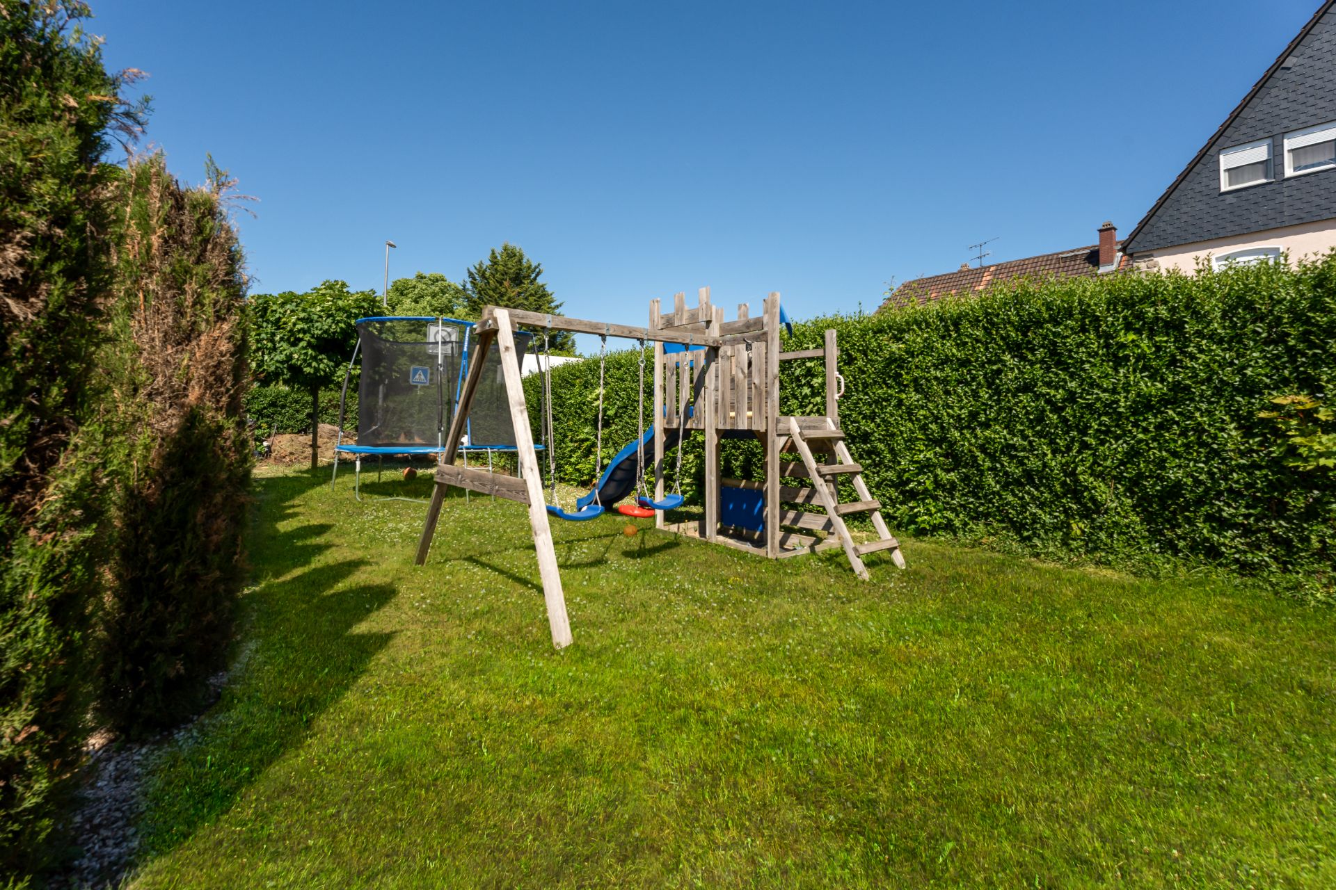 Garten mit Spielplatz: Holzspielturm, Schaukel und Trampolin auf Rasen.