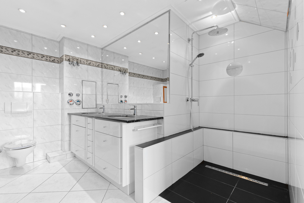 Modernes, weiß gefliestes Badezimmer mit Doppelwaschbecken, großer Dusche und Wandtoilette.