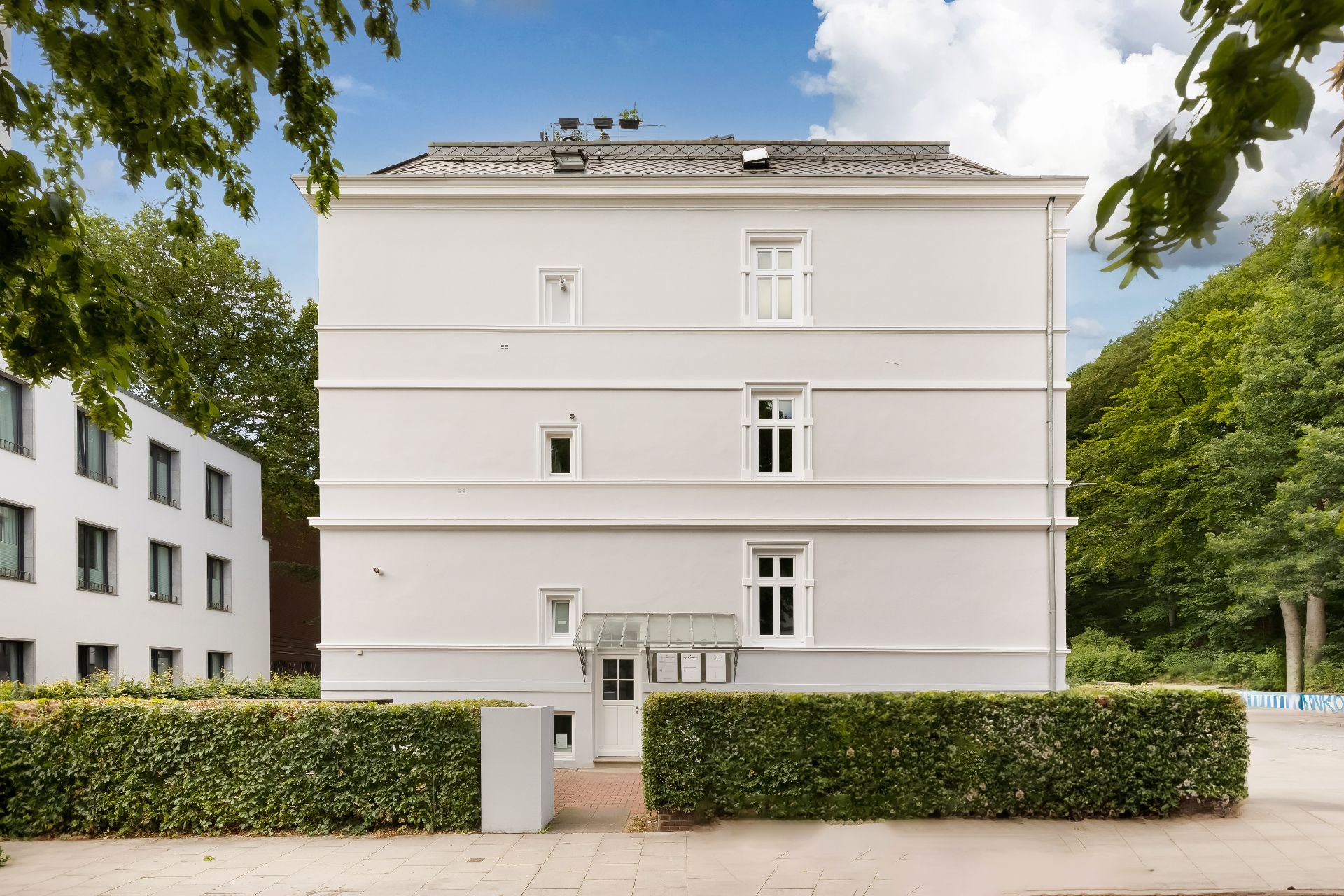 Weiße Wohnhausfassade mit mehreren Fenstern, Büschen und Gehweg im Vordergrund.