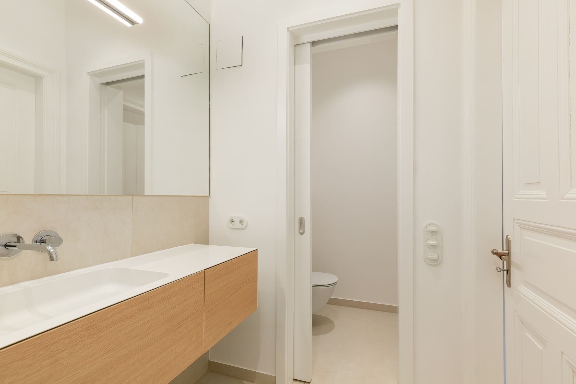Modernes Badezimmer mit großem Spiegel, Waschbecken und teilweise sichtbarem WC hinter einer Schiebetür.