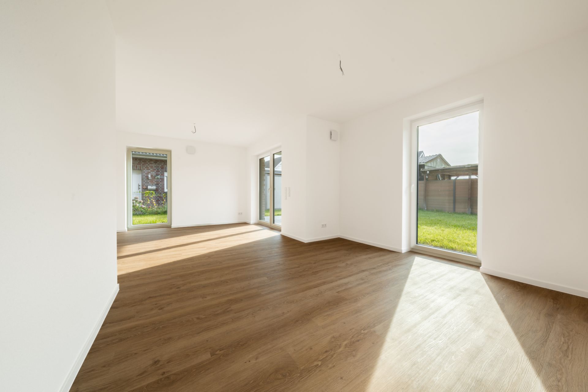 Heller, leerer Raum mit Holzfußboden und großen Fenstern zum Garten.