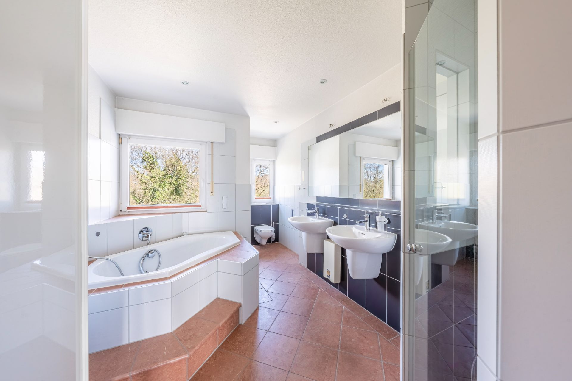 Helles Badezimmer mit Eckbadewanne, zwei Waschbecken, Spiegel und zwei Fenstern.