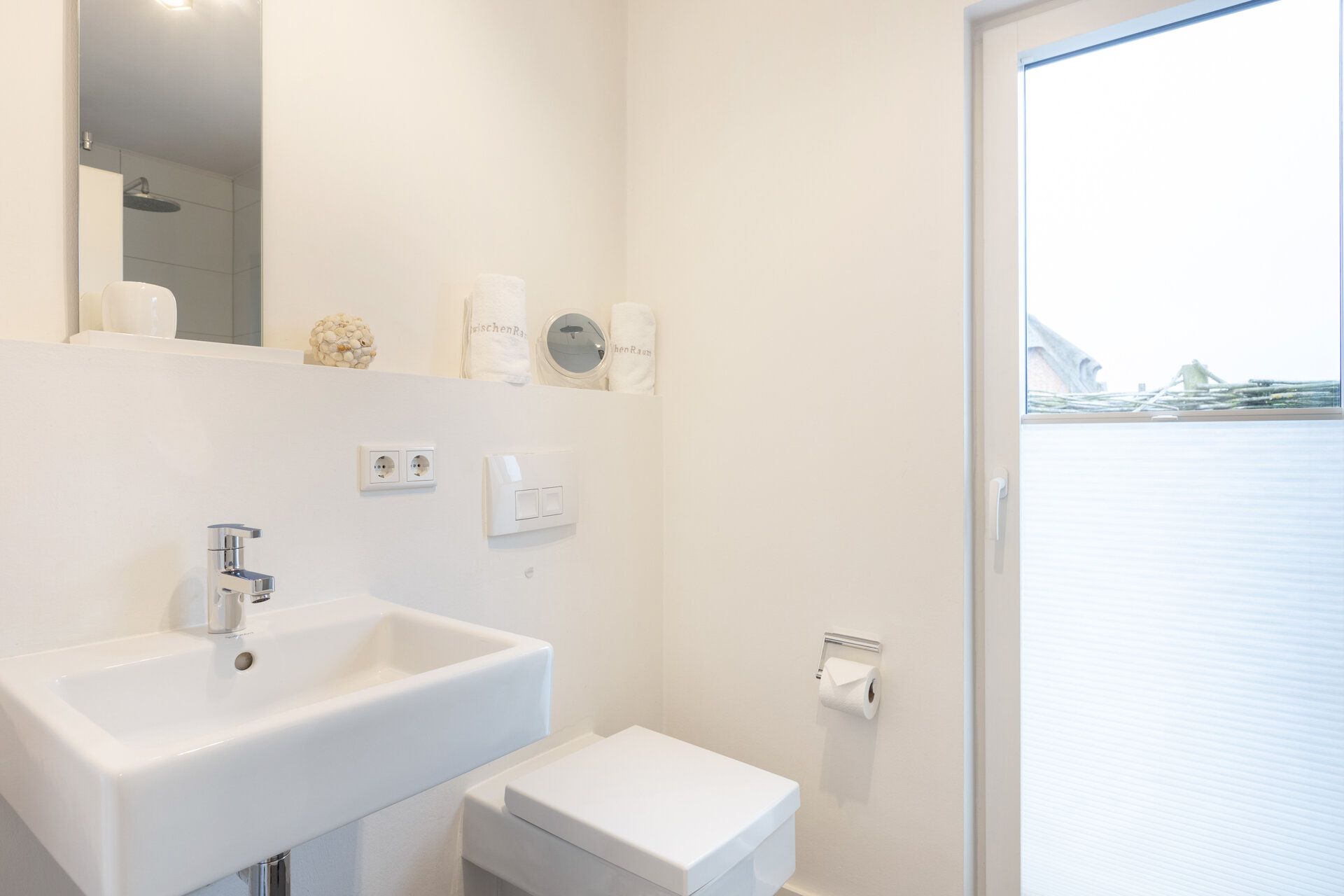 Helles Badezimmer mit Waschbecken, WC, Spiegel und Fenster mit Sichtschutz.