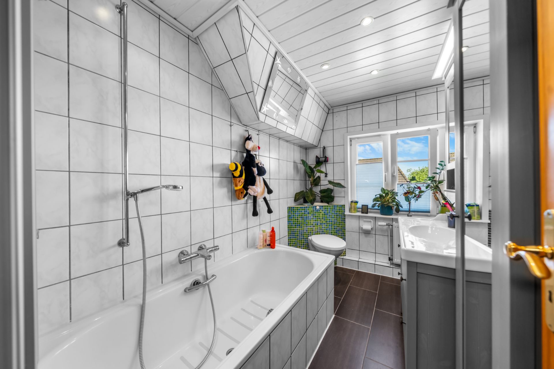 Modernes Badezimmer mit Badewanne, Fenster, Pflanzen und weißen Fliesen an Wand und Decke.