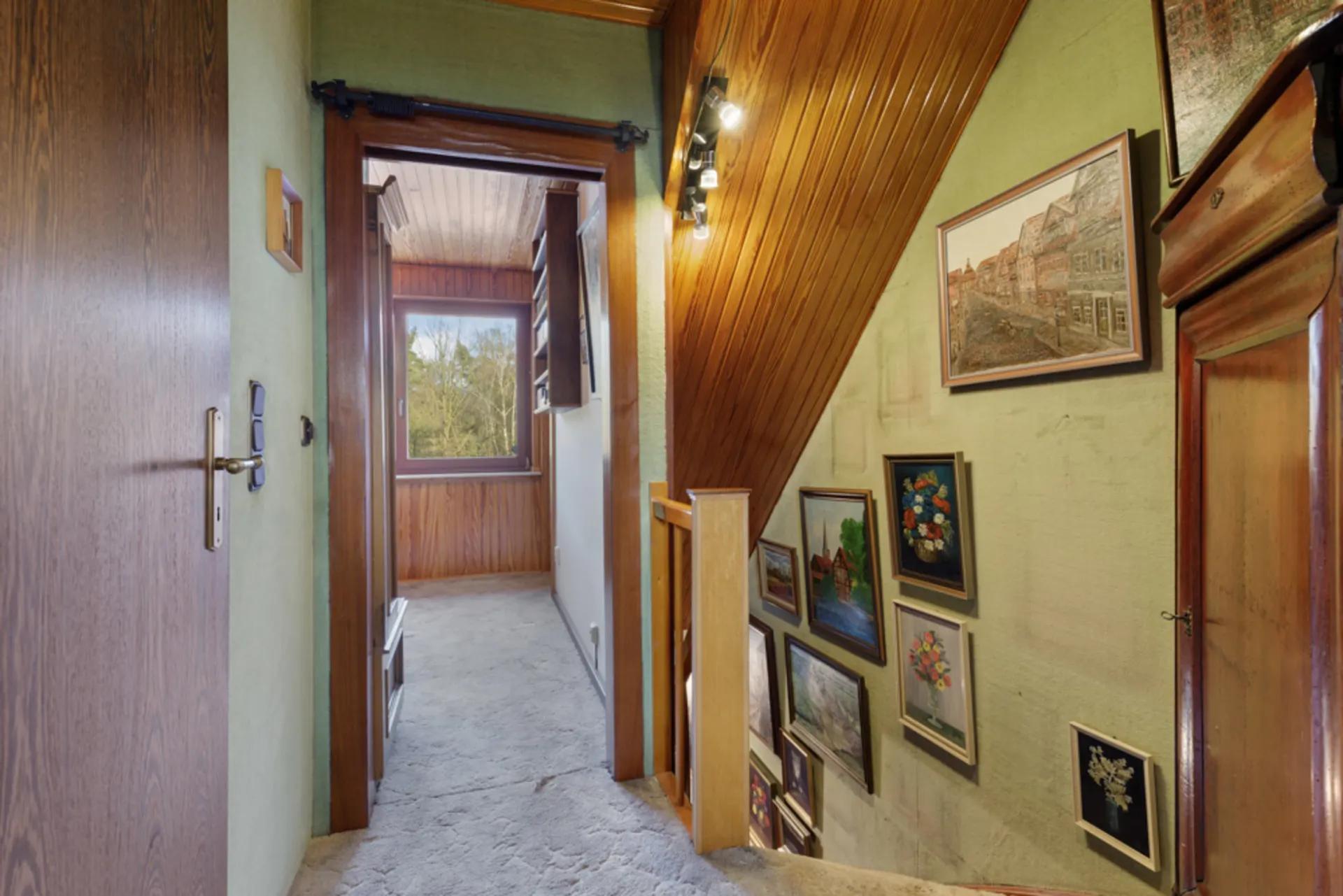 Flur mit Teppichboden, Holzwänden und mehreren Bildern an der Wand, Fenster mit Blick ins Grüne.