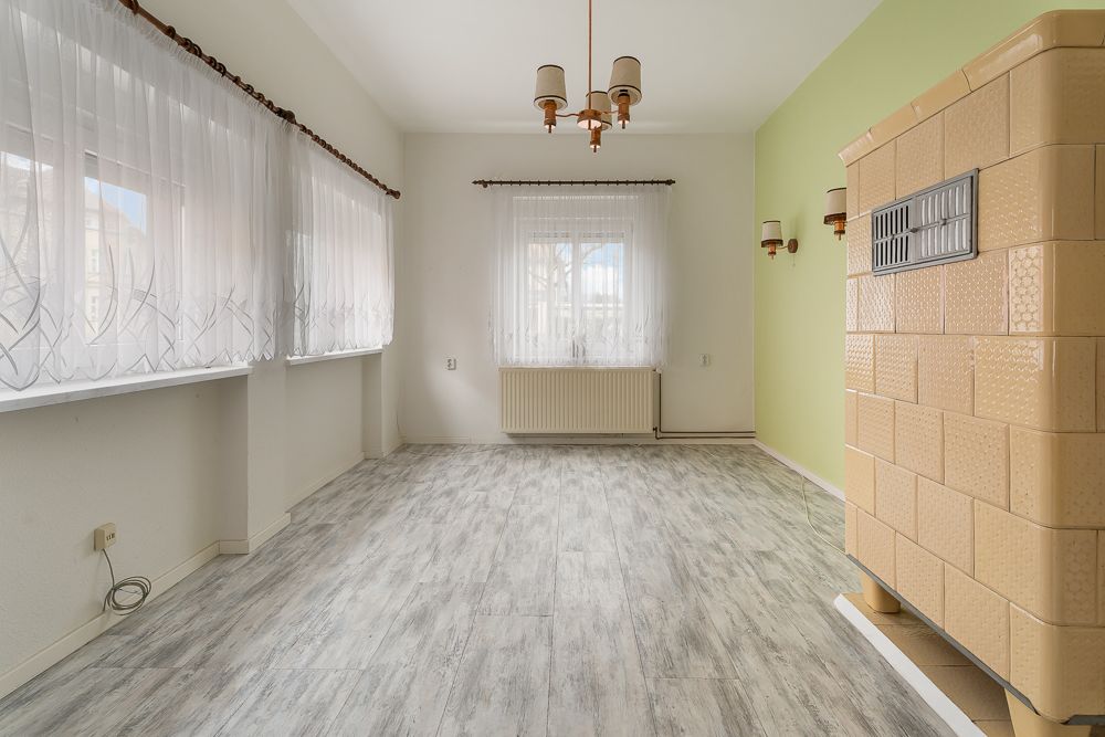 Helles Zimmer mit großen Fenstern, hellem Boden und einem Kachelofen an der rechten Wand.