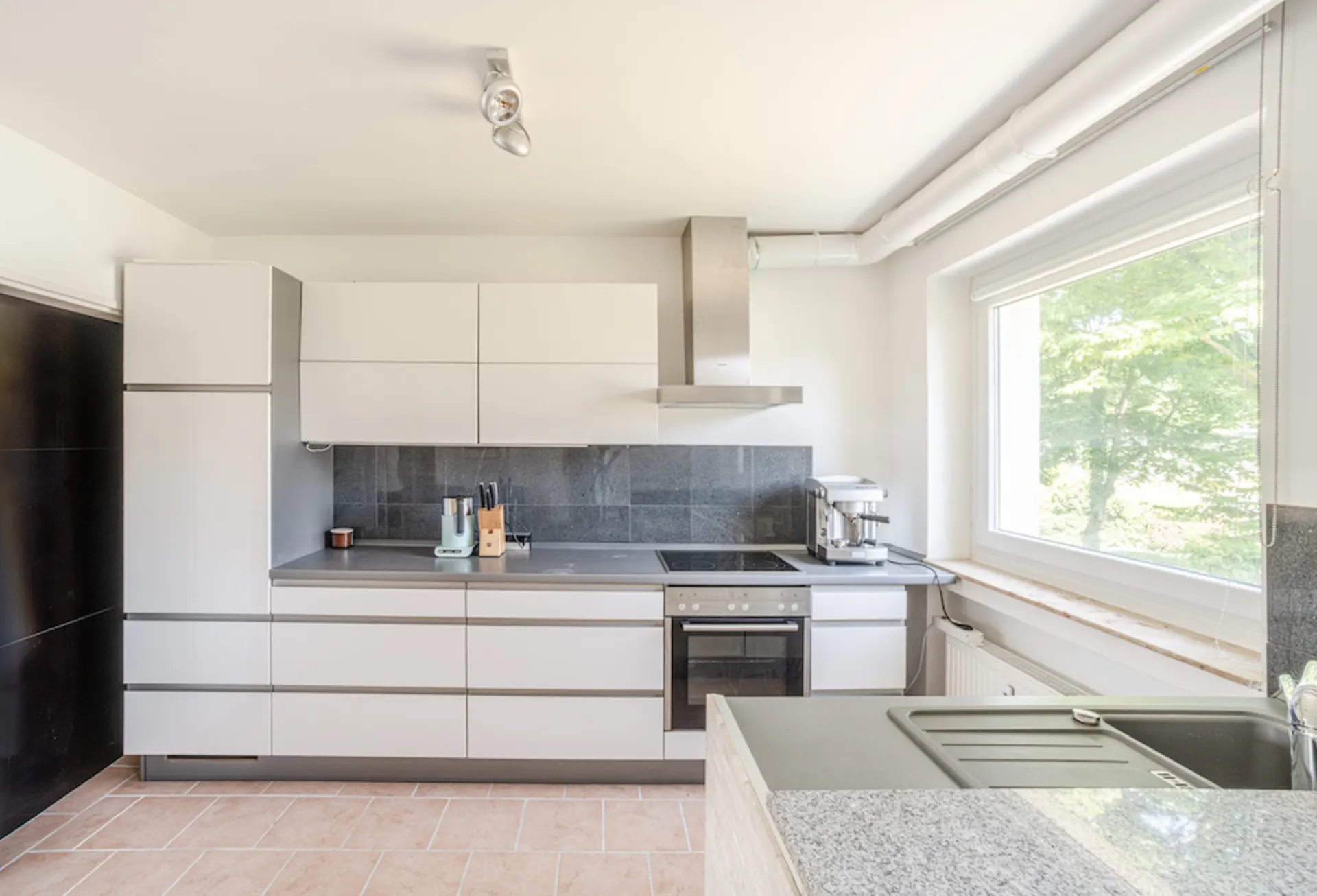 Moderne weiße Einbauküche mit Elektrogeräten, großem Fenster und grauer Arbeitsplatte.
