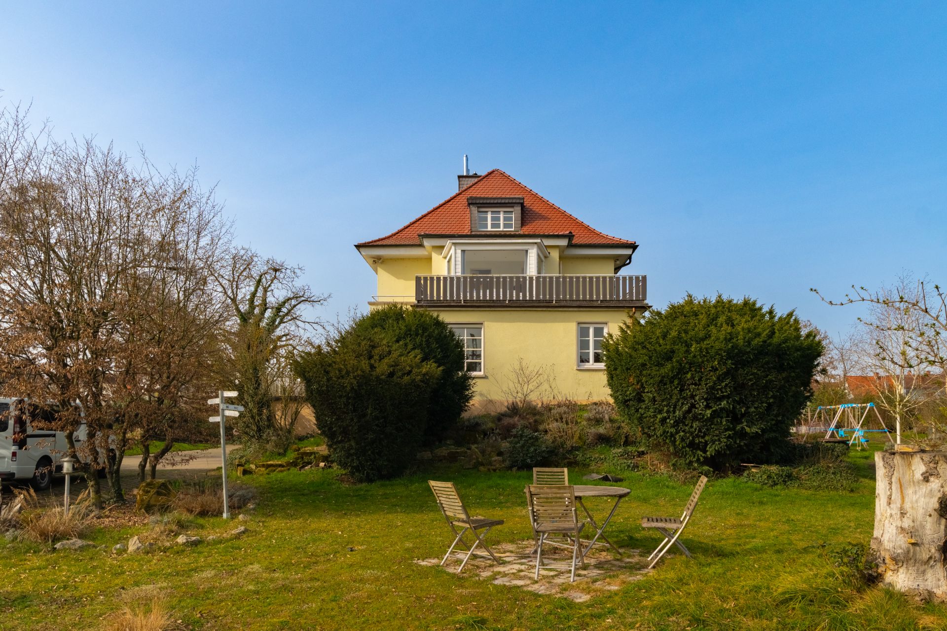 Gelbes Haus mit rotem Dach, Garten, Gartenmöbeln und Bäumen im Vordergrund.