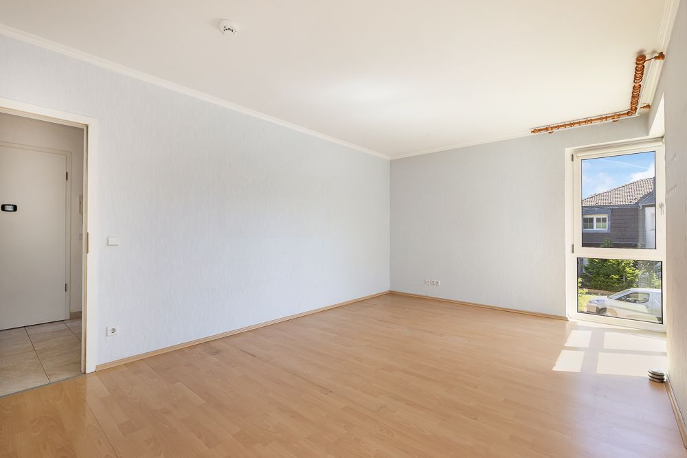 Heller, leerer Raum mit Holzfußboden, weißer Wand und großem Fenster.
