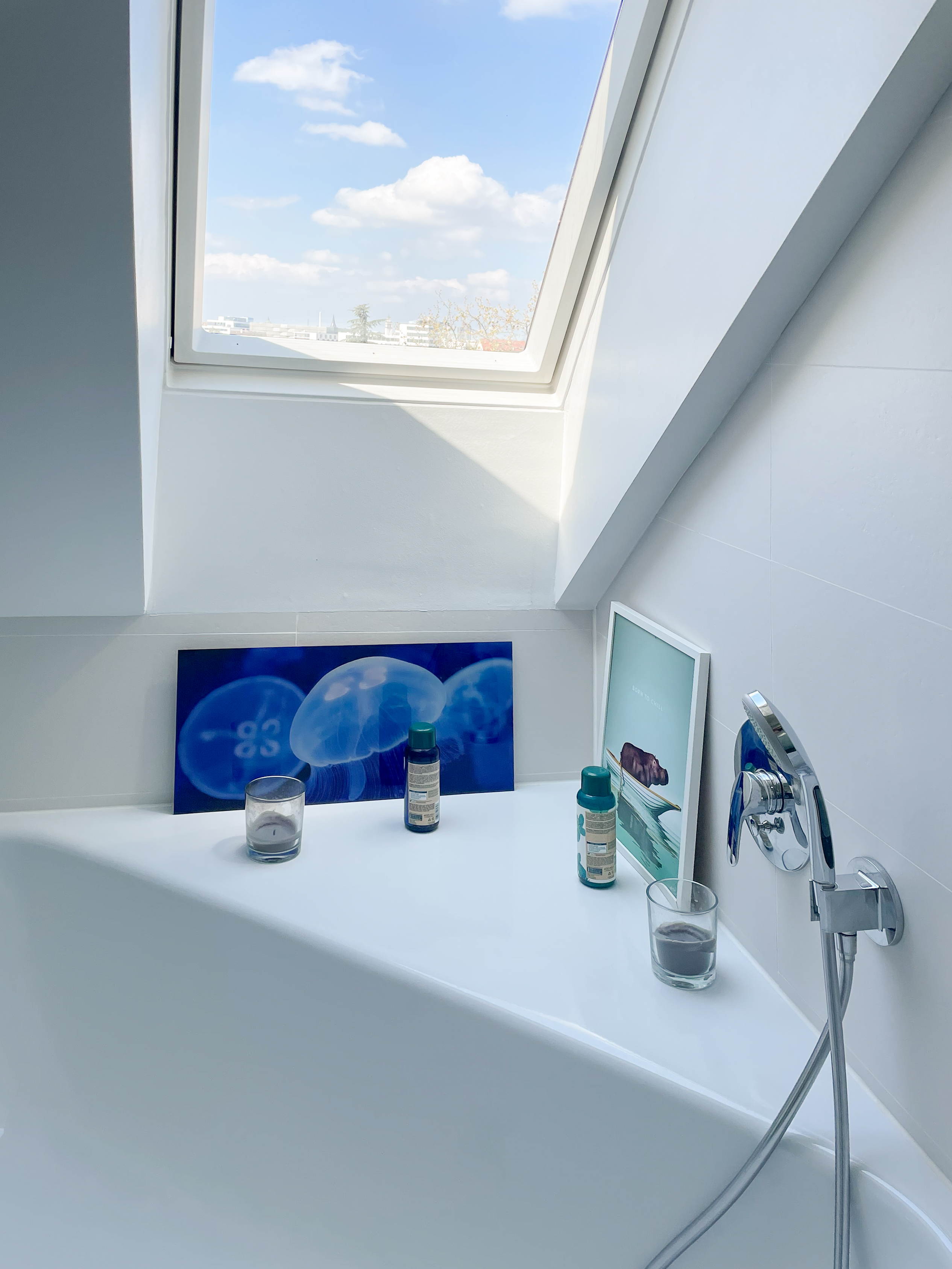 Badewanne unter einem Fenster mit Bildern, Kerzen und Kosmetikflaschen auf dem Rand.
