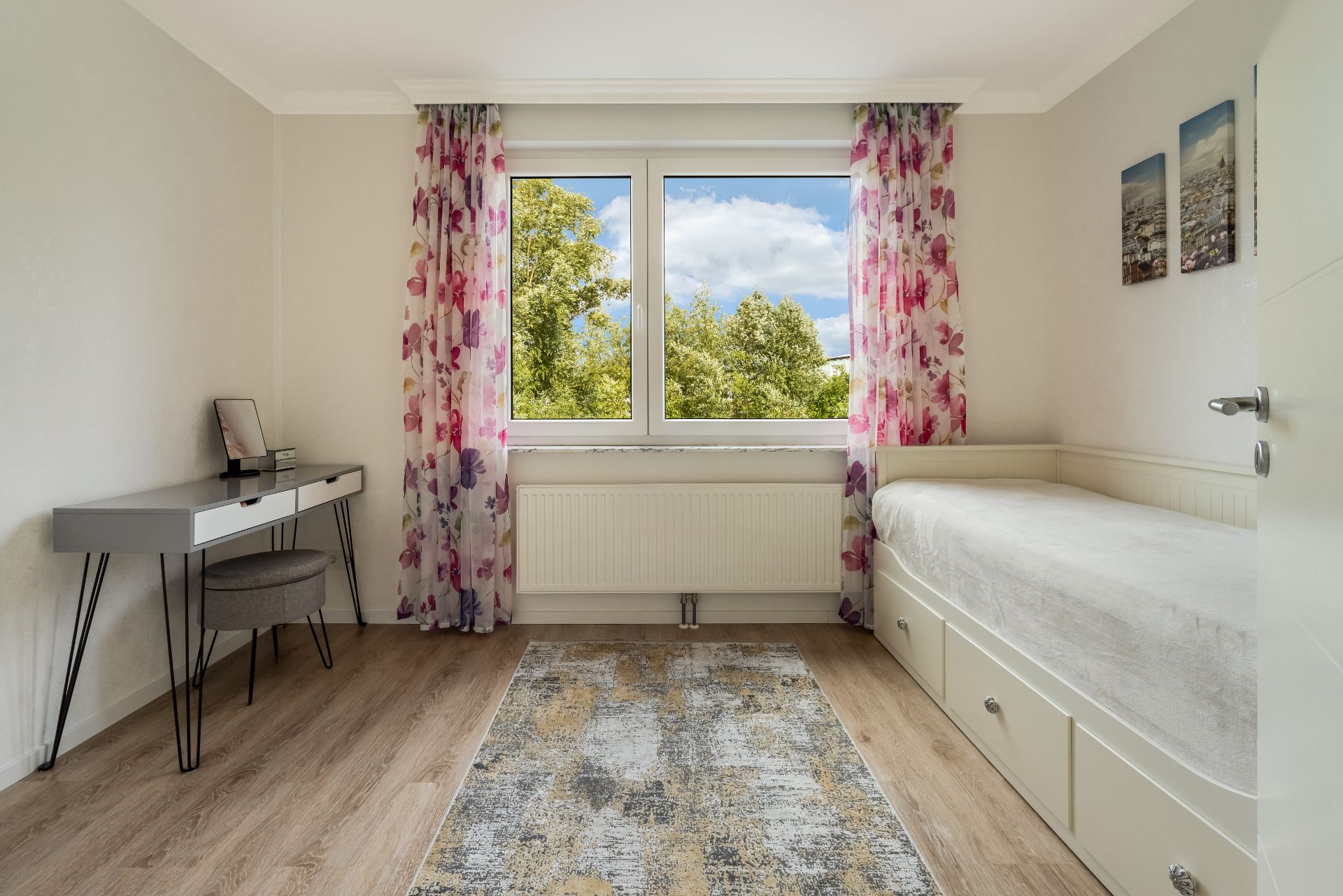 Helles Zimmer mit Einzelbett, Schreibtisch, bunten Vorhängen und großem Fenster mit Ausblick ins Grüne.