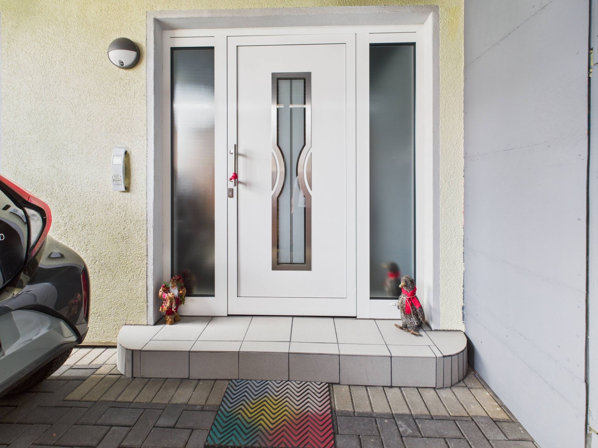 Weiße Haustür mit zwei Seitenscheiben, bunte Fußmatte und zwei Dekofiguren auf den Stufen.