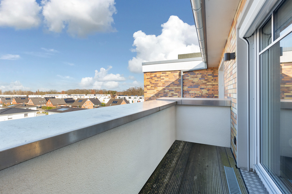 Balkon mit Holzboden und Blick auf Wohnhäuser und blauen Himmel mit Wolken.