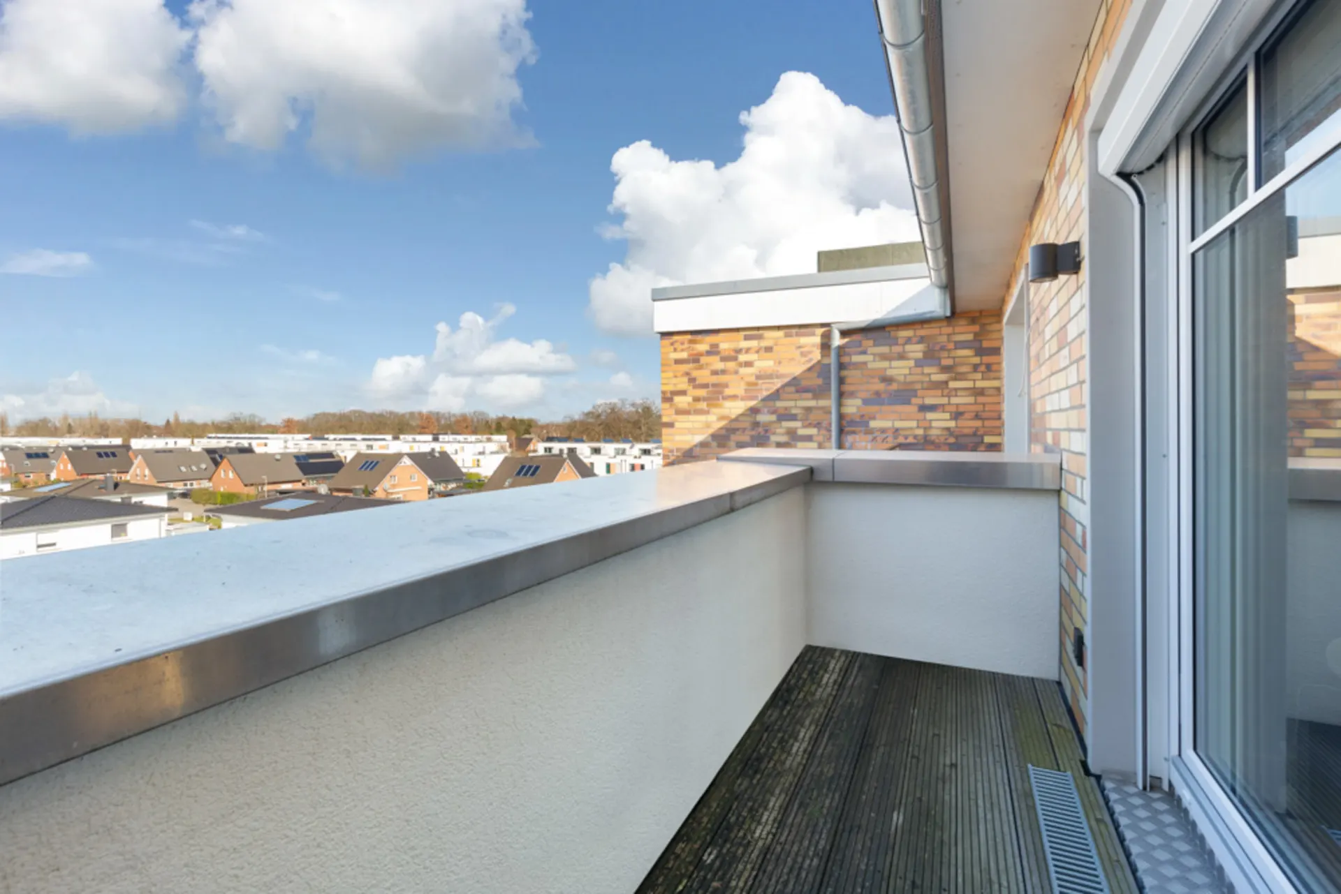 Balkon mit Holzboden und Blick auf Wohnhäuser und blauen Himmel mit Wolken.