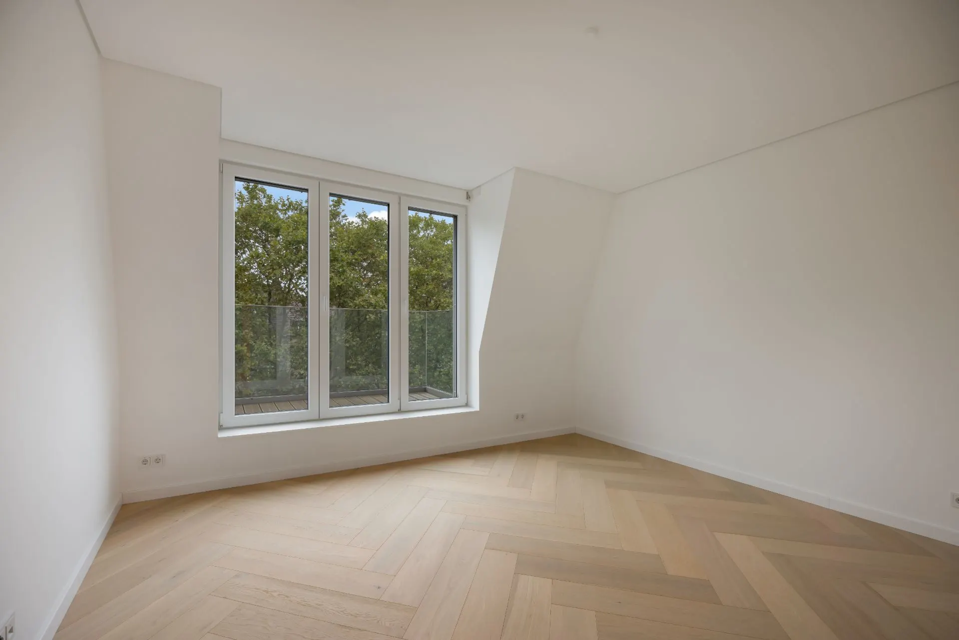 Helles, leeres Zimmer mit großem Fenster und Parkettboden.