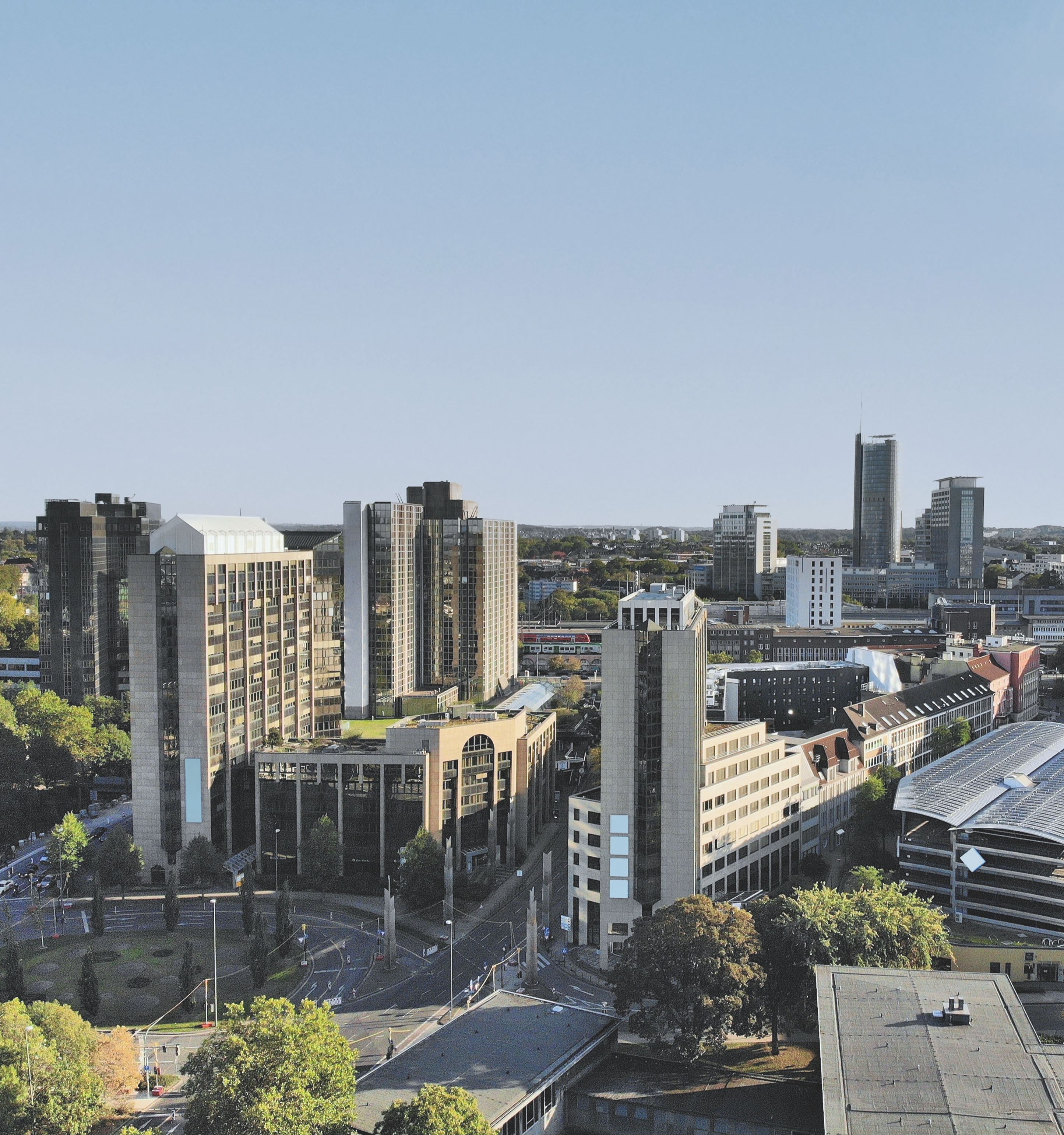 Stadtansicht mit mehreren modernen Hochhäusern und Bäumen im Vordergrund.