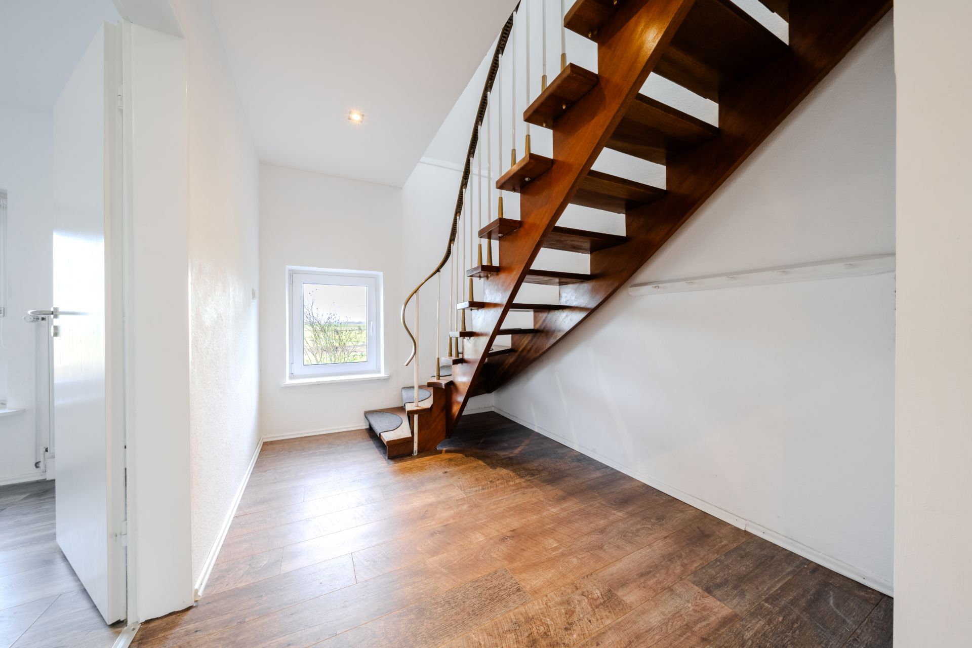 Innenraum mit Holztreppe, weißer Wand und Fenster, Holzboden.