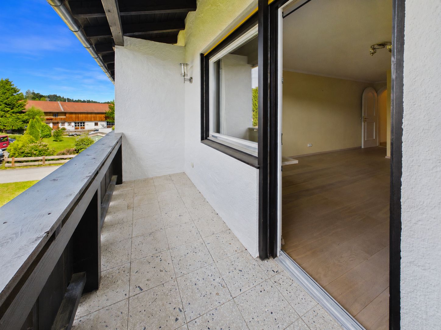 Balkon mit Fliesenboden an einem Wohnhaus, daneben ein Raum mit Holzboden und offenem Fenster.