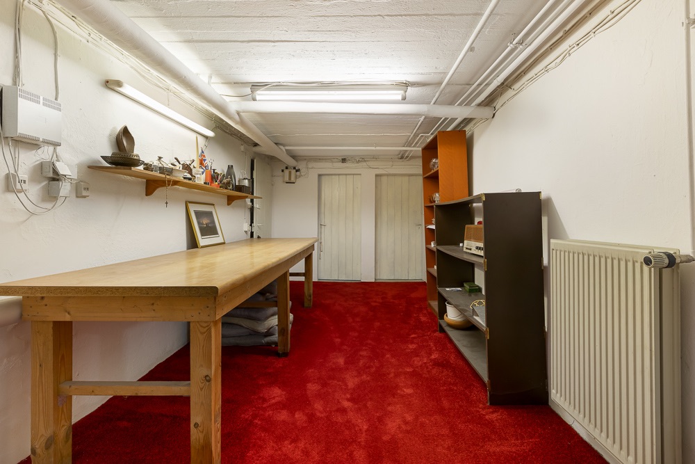 Kellerraum mit rotem Teppich, Holztisch, Regal und Heizkörper an der Wand.
