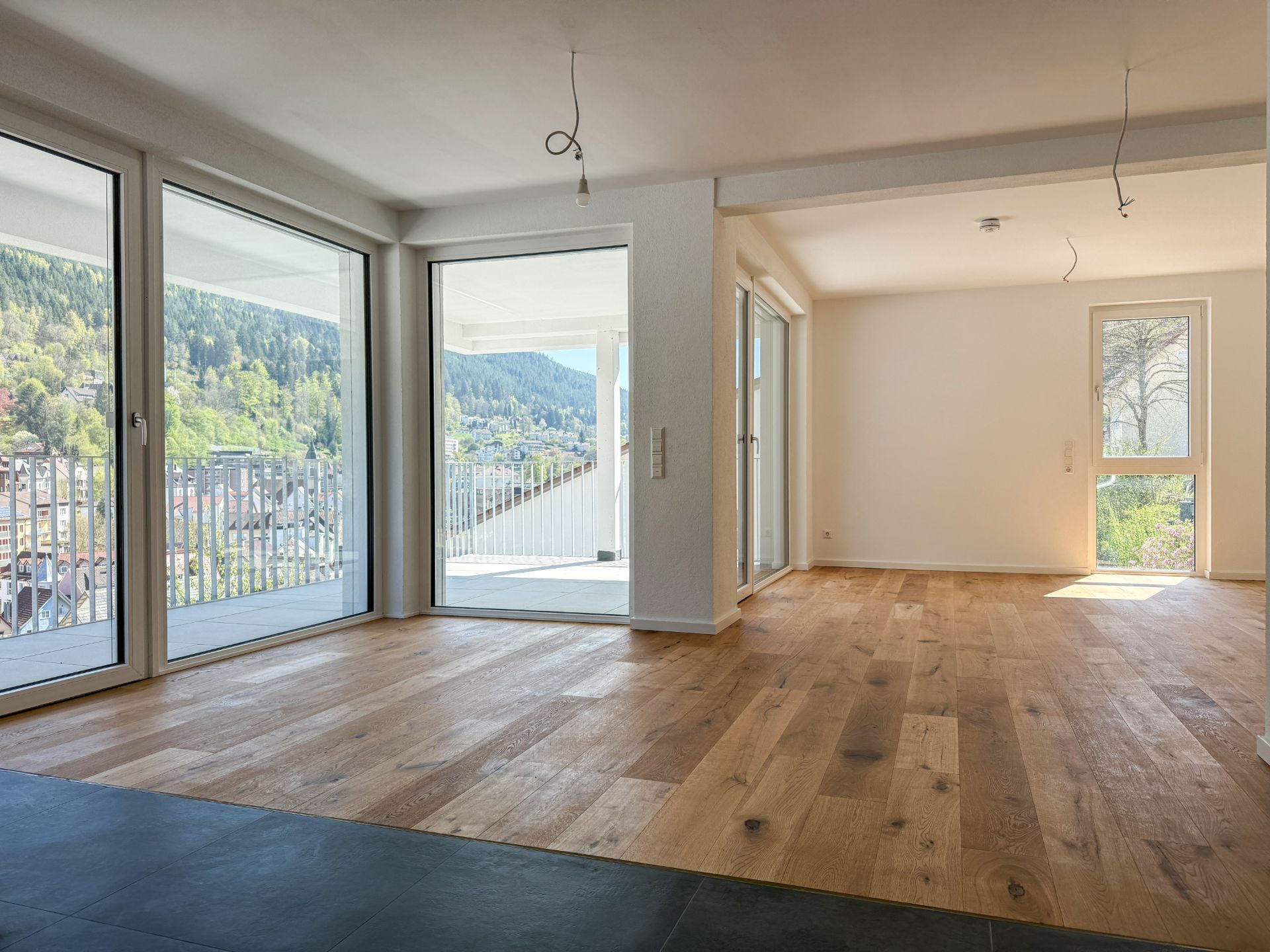 Heller Wohnraum mit Holzfußboden, großen Fenstern und Blick auf einen Balkon und grüne Hügel.