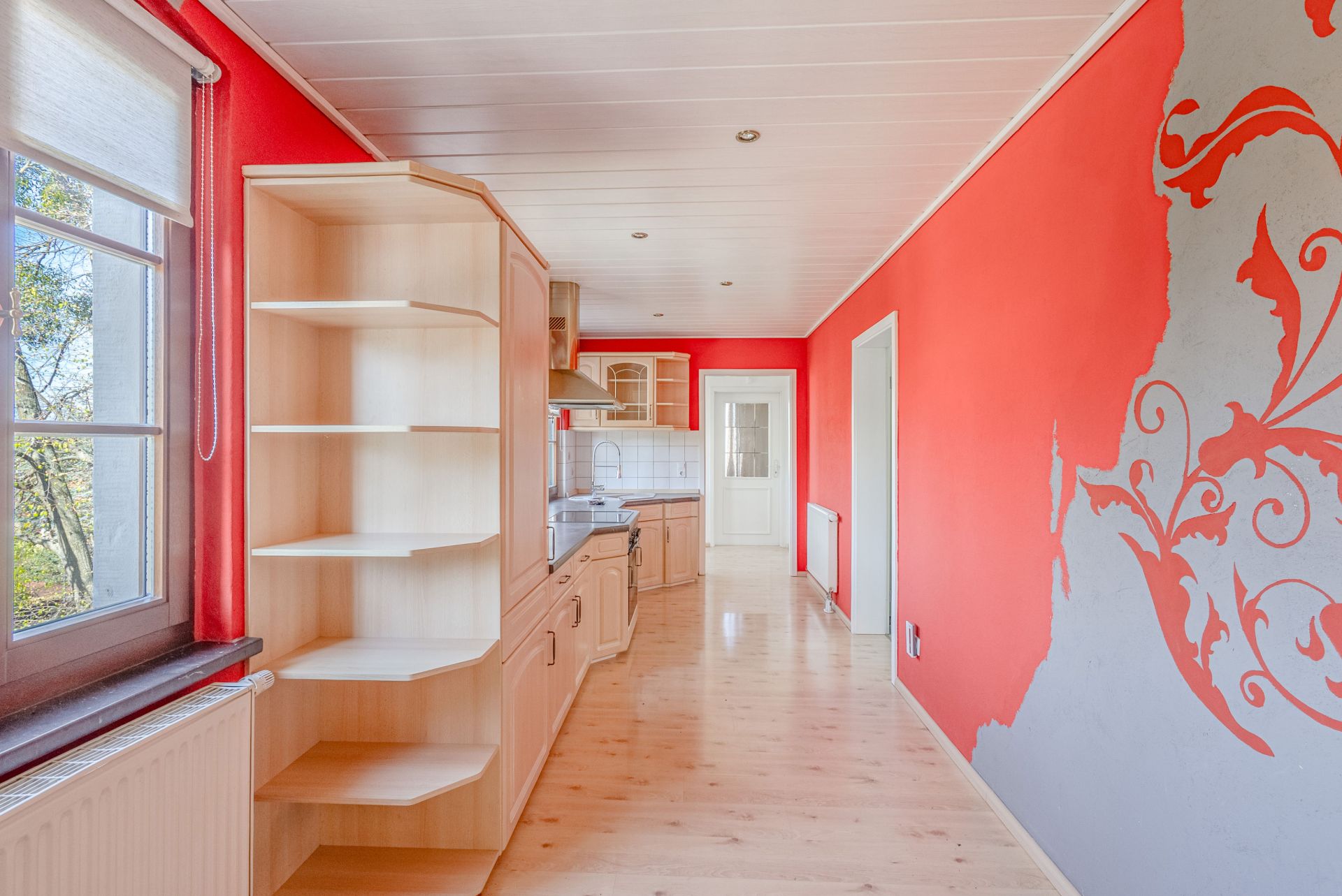 Lange Küche mit roter Wand, Holzregalen, großem Fenster und gemustertem Wanddekor.