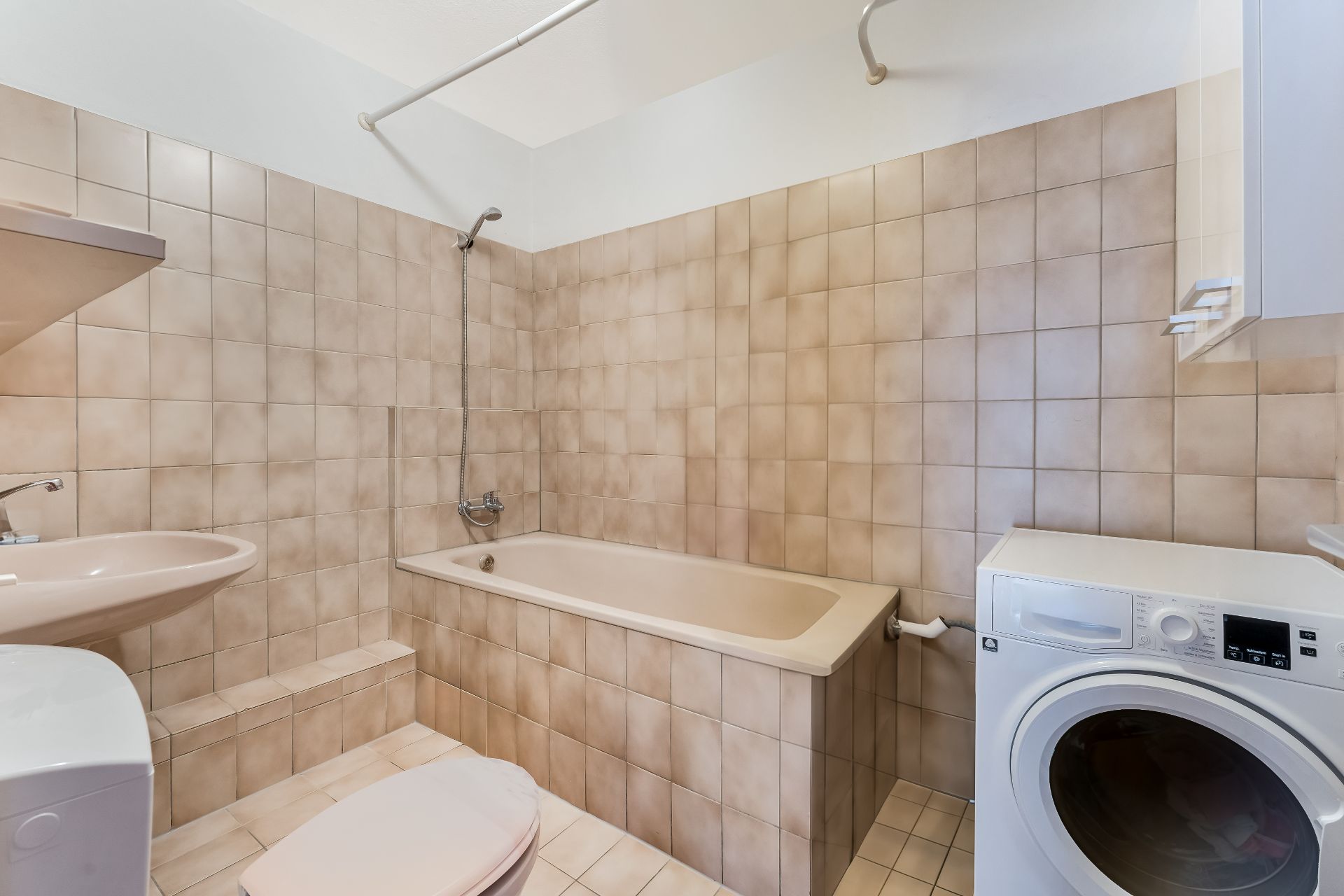 Badezimmer mit Badewanne, Waschbecken, WC und Waschmaschine, beige Fliesen an Wand und Boden.