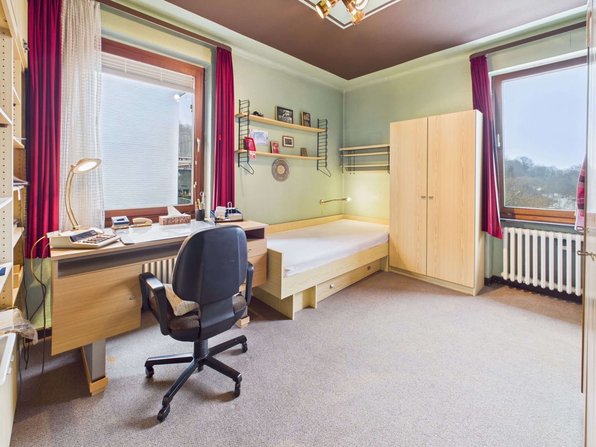 Helles Schlafzimmer mit Schreibtisch, Stuhl, Bett, Kleiderschrank und zwei Fenstern mit roten Vorhängen.