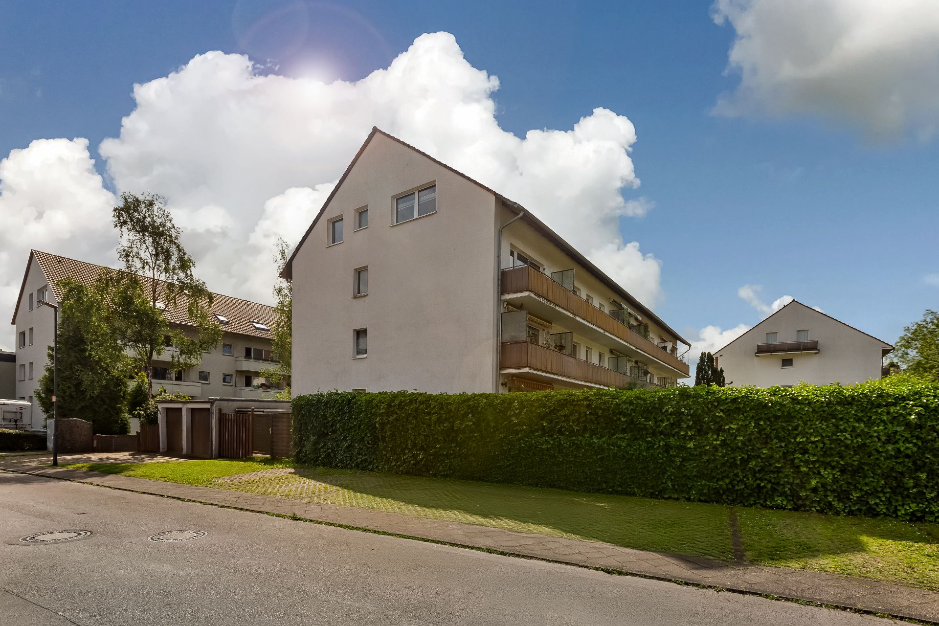 Mehrfamilienhaus mit Balkonen, Hecke und Garagen an einer ruhigen Straße bei Sonnenschein.