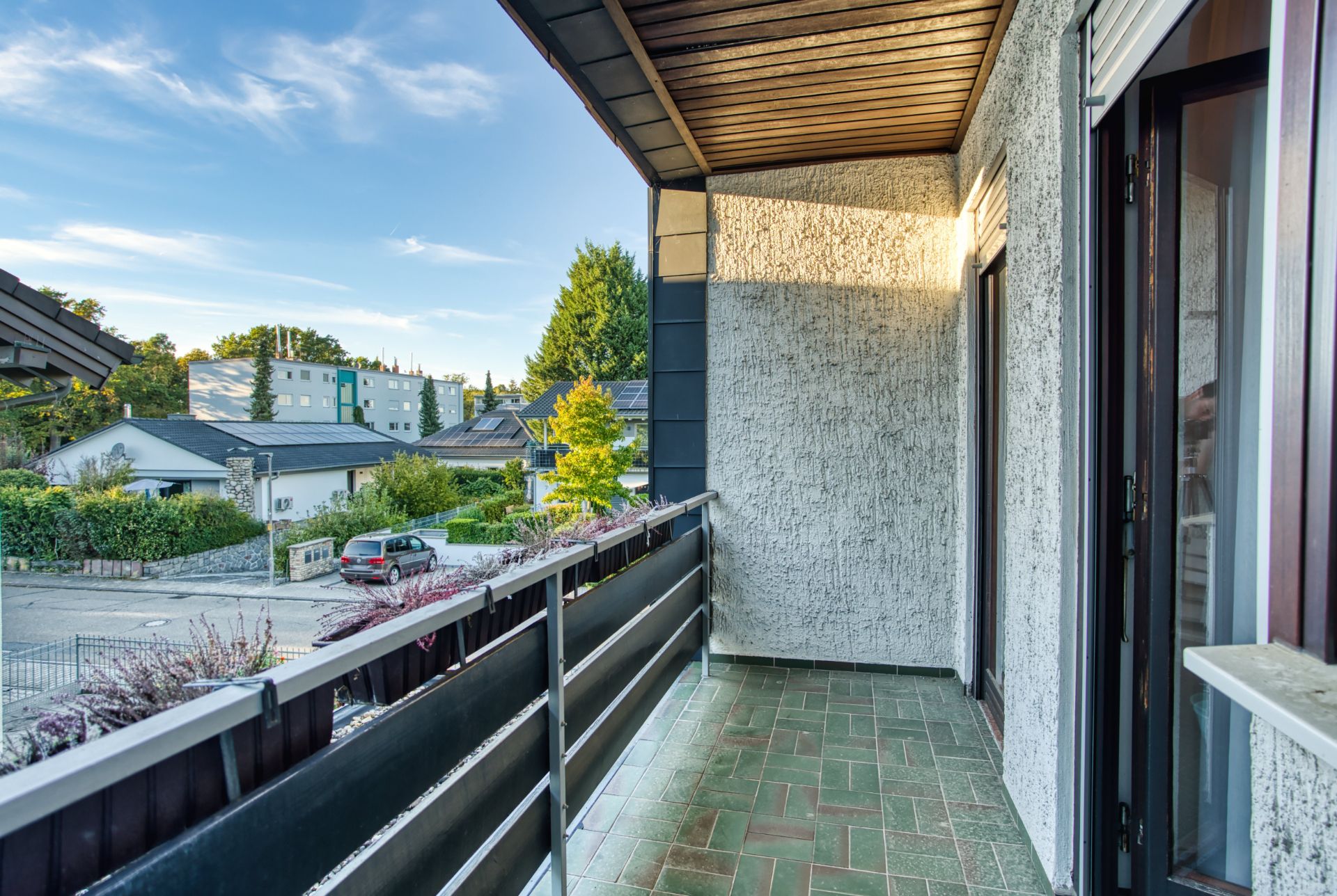 Balkon mit grün gefliestem Boden und Metallgeländer, Ausblick auf Wohngegend mit Bäumen.