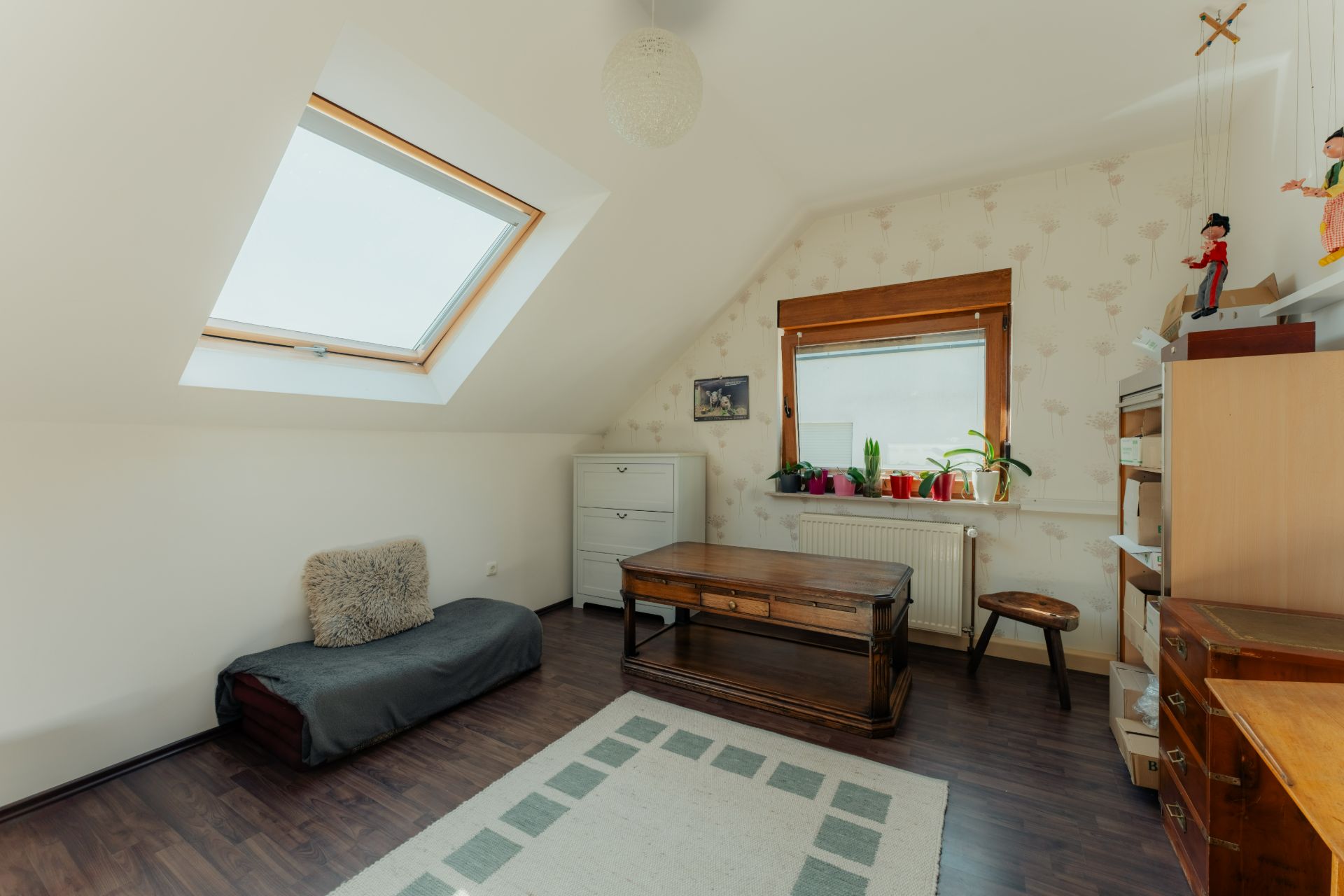 Helles Zimmer mit Dachfenster, Holzboden, Sofa, Tisch, Regal und Pflanzen am Fenster.
