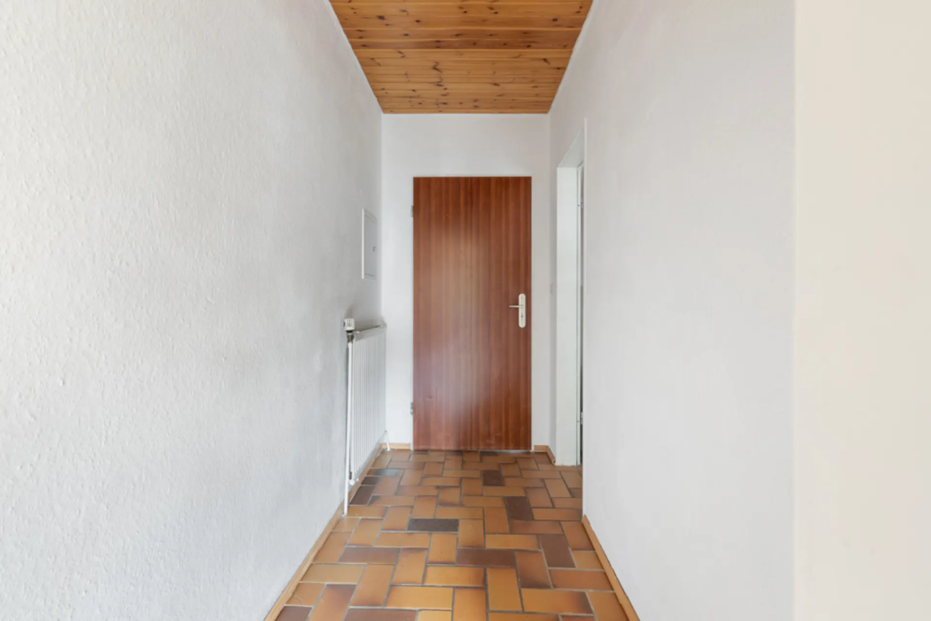 Schmaler Flur mit braunen Fliesen, Holztür, Holzdecke und weiß gestrichenen Wänden.
