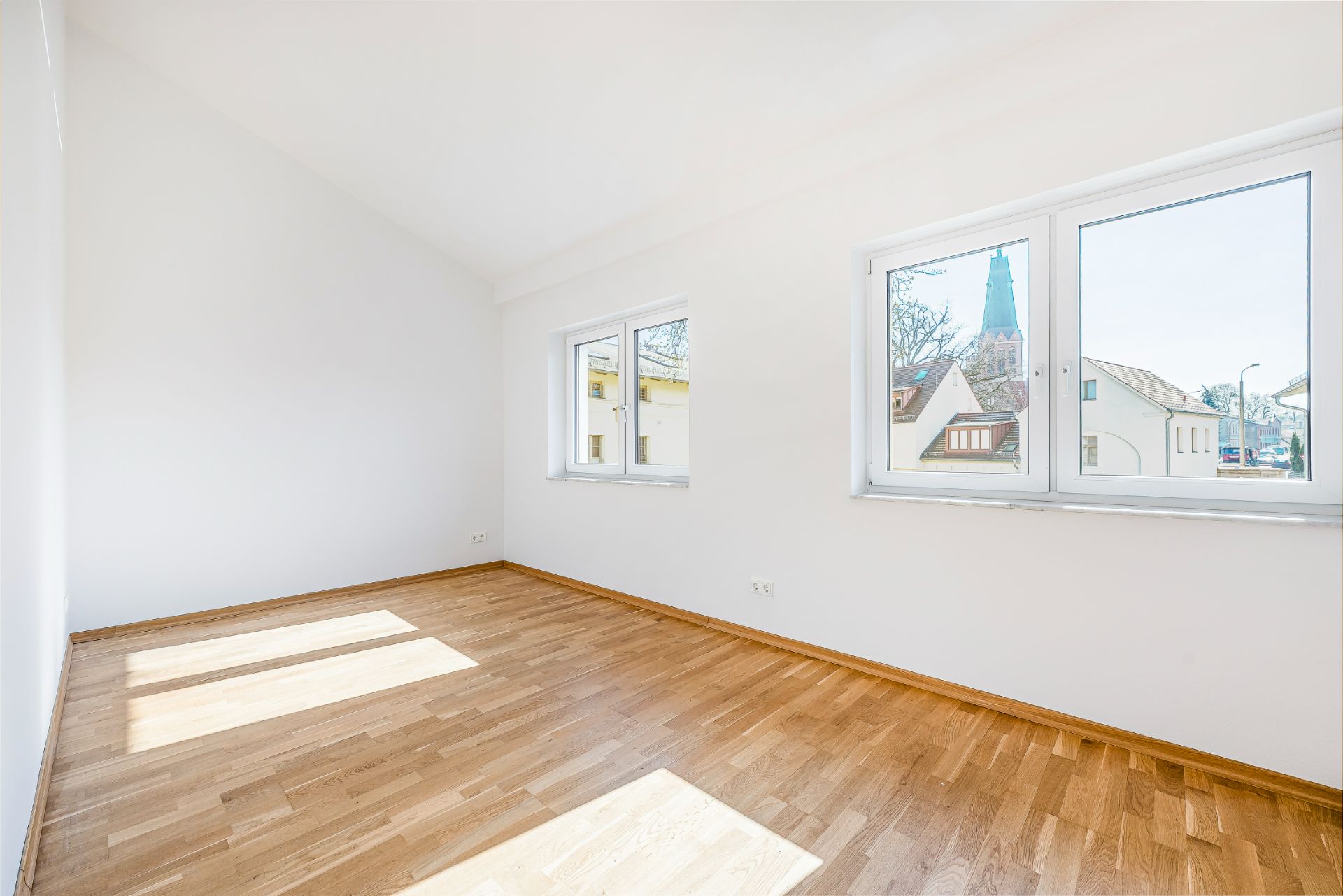 Helles Zimmer mit weiß gestrichenen Wänden, Parkettboden und drei Fenstern, durch die Tageslicht fällt.
