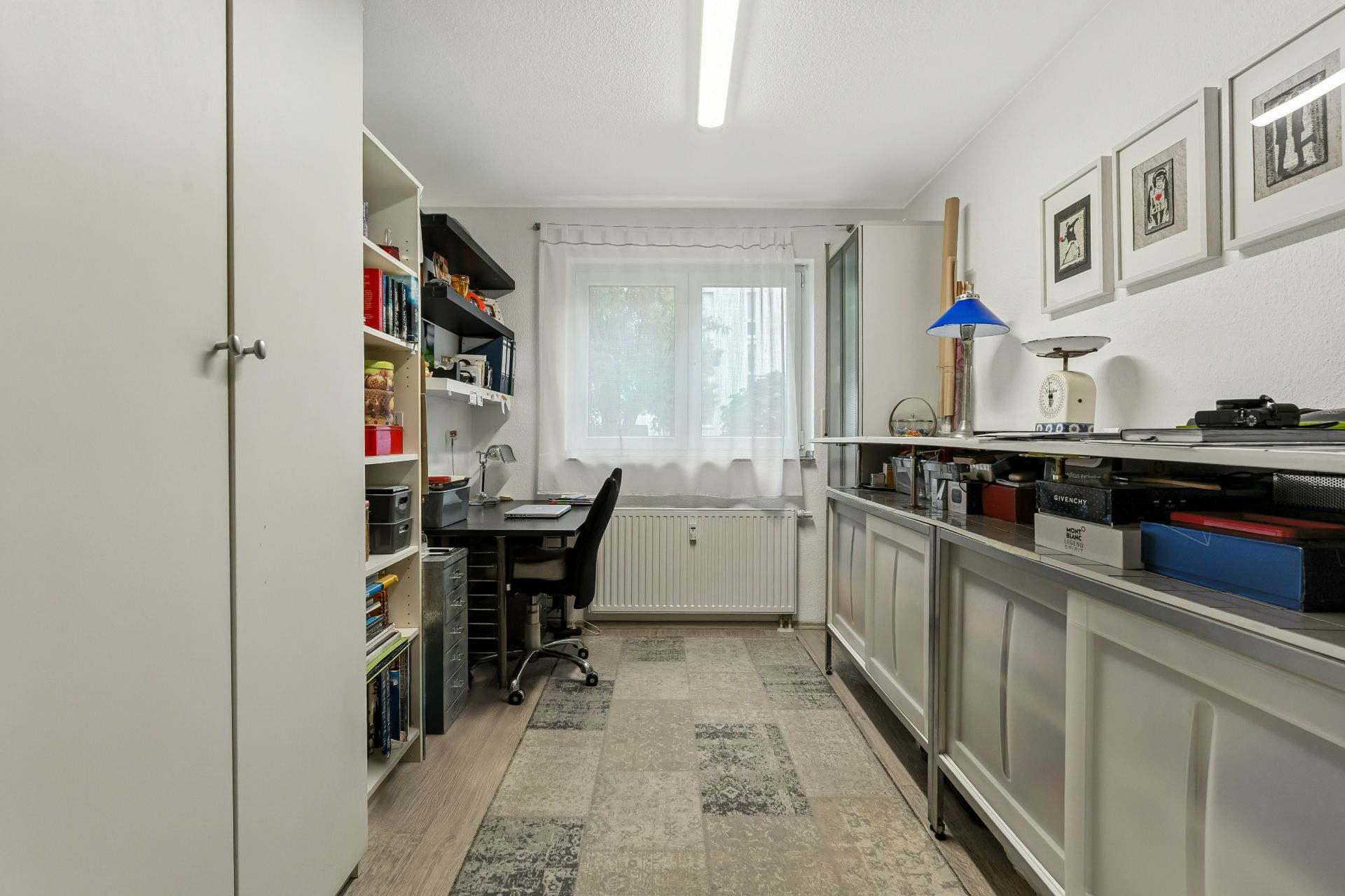 Helles Arbeitszimmer mit Schreibtisch, Regal, Fenster und Bildern an der Wand.