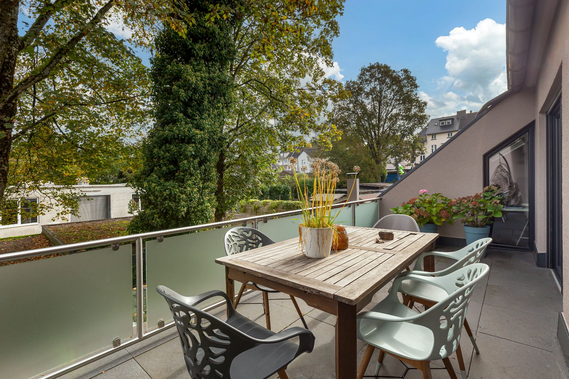 Balkon mit Holztisch, sechs Stühlen, Pflanzen und Blick auf Bäume und Nachbarhäuser.