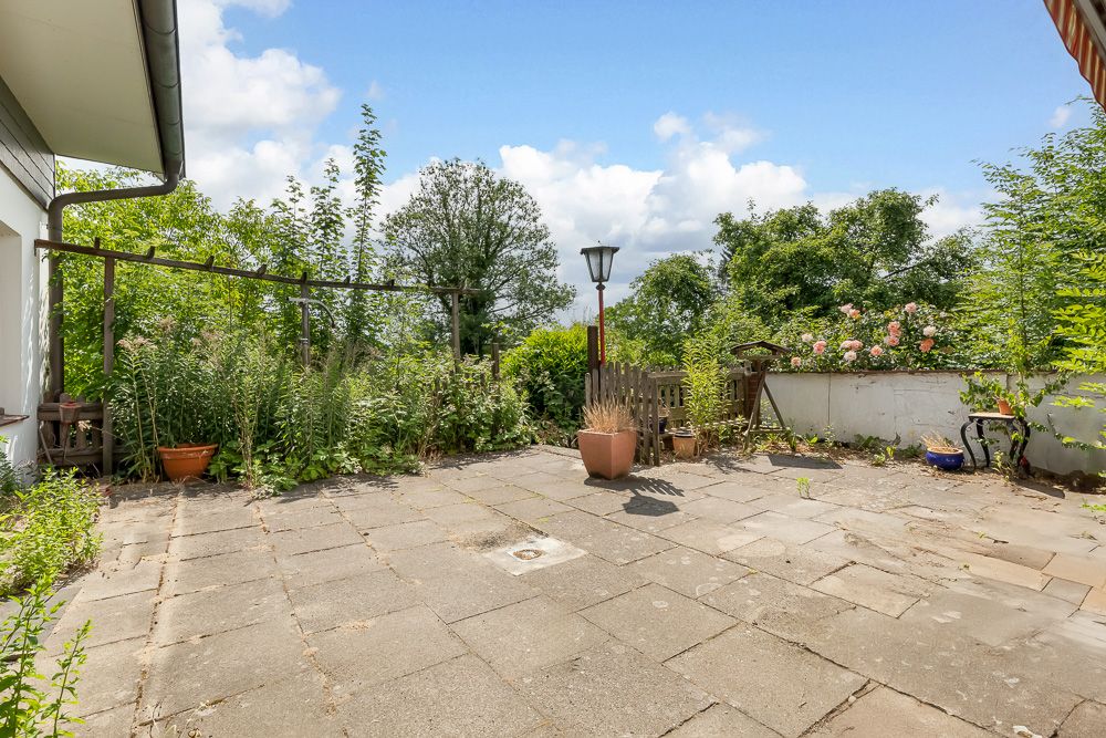 Steinplatten-Terrasse mit Pflanztöpfen, Laterne und Bewuchs am Rand, umgeben von Bäumen.