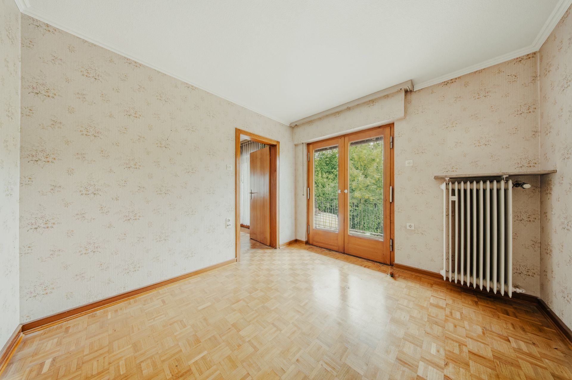 Leerer Raum mit Parkettboden, Tapeten, großem Fenster, Heizkörper und Tür zum Balkon.