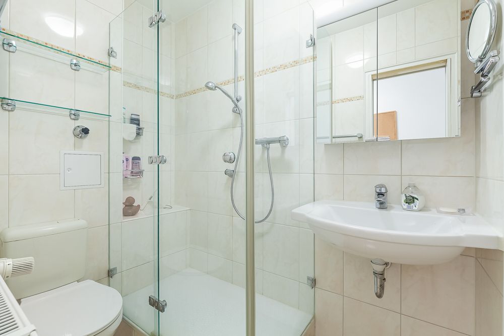 Helles Bad mit Dusche, Toilette, Waschbecken und Spiegel, weiße Fliesen an den Wänden.