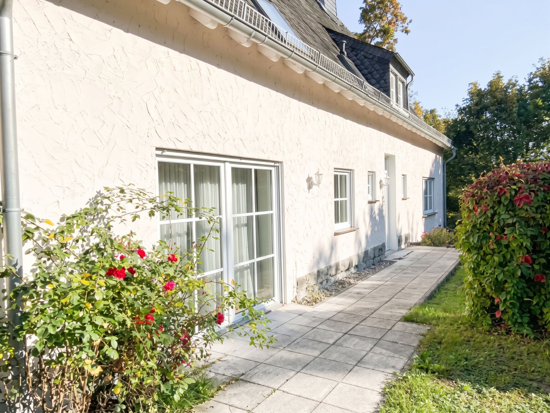 Heller Hauszugang mit großen Fenstern, Blumenbeeten und gepflastertem Weg im Garten.