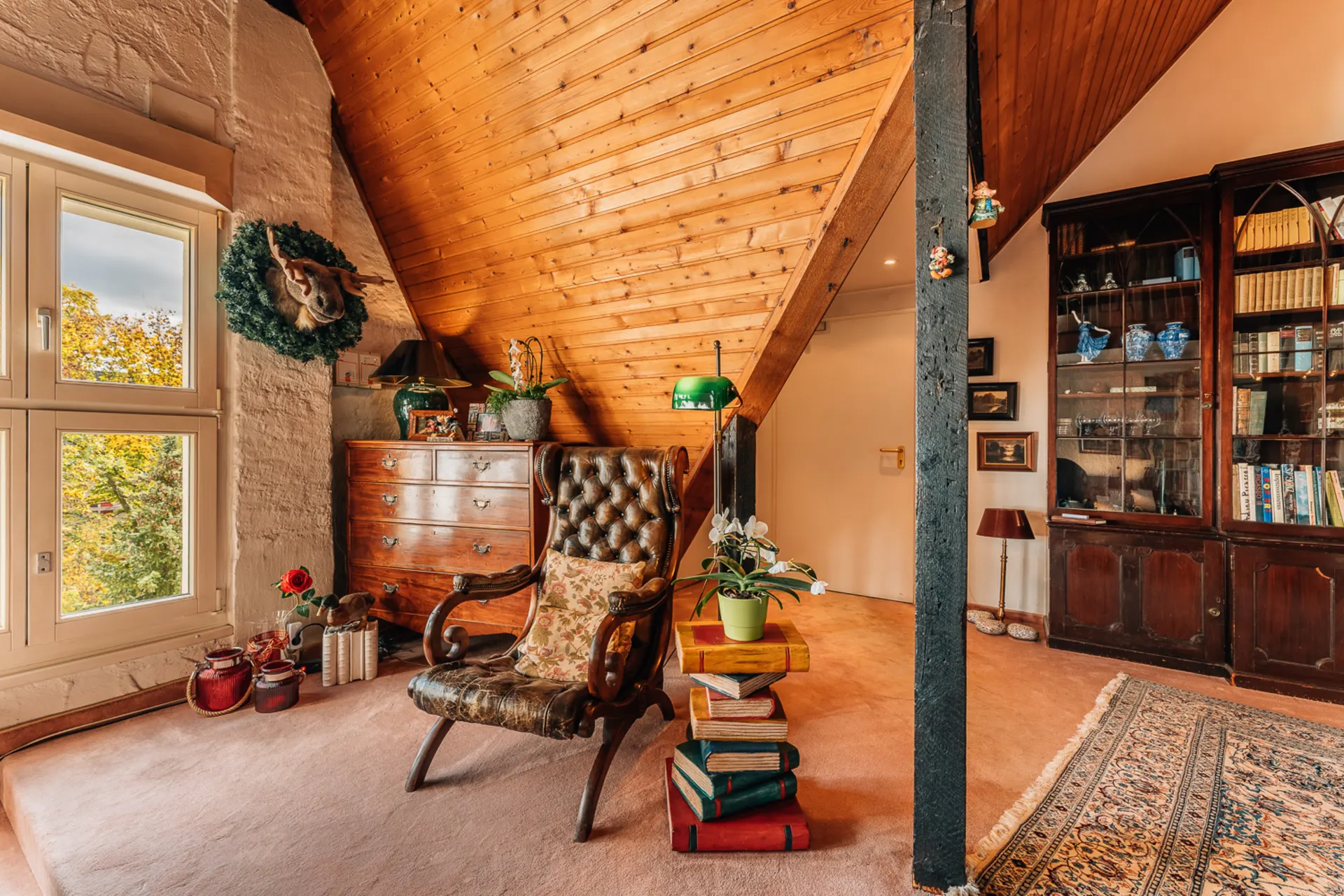 Gemütlicher Dachwohnraum mit Sessel, Holzmöbeln, Bücherregal und Fenstern mit Blick ins Grüne.