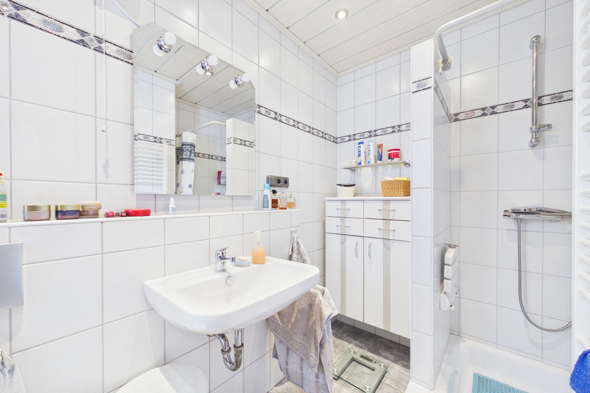 Helles Badezimmer mit Waschbecken, Spiegel, Dusche und weißem Schrank auf Fliesenboden.
