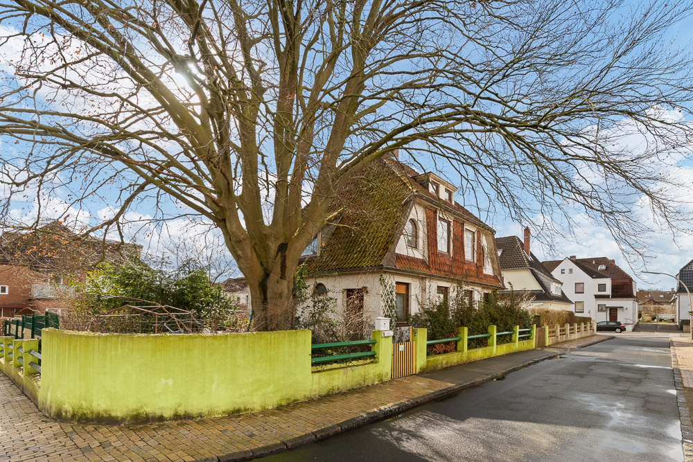 Zweistöckiges Wohnhaus mit grünem Zaun und großem Baum an einer ruhigen Straße.