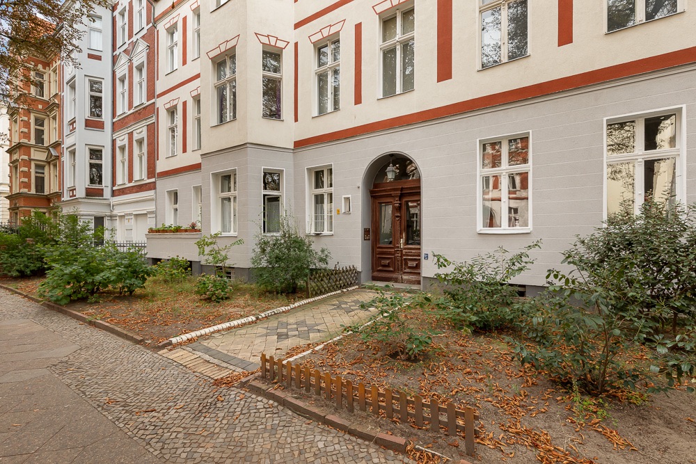 Eingang eines Mehrfamilienhauses mit Vorgarten und Gehweg, umgeben von Pflanzen.
