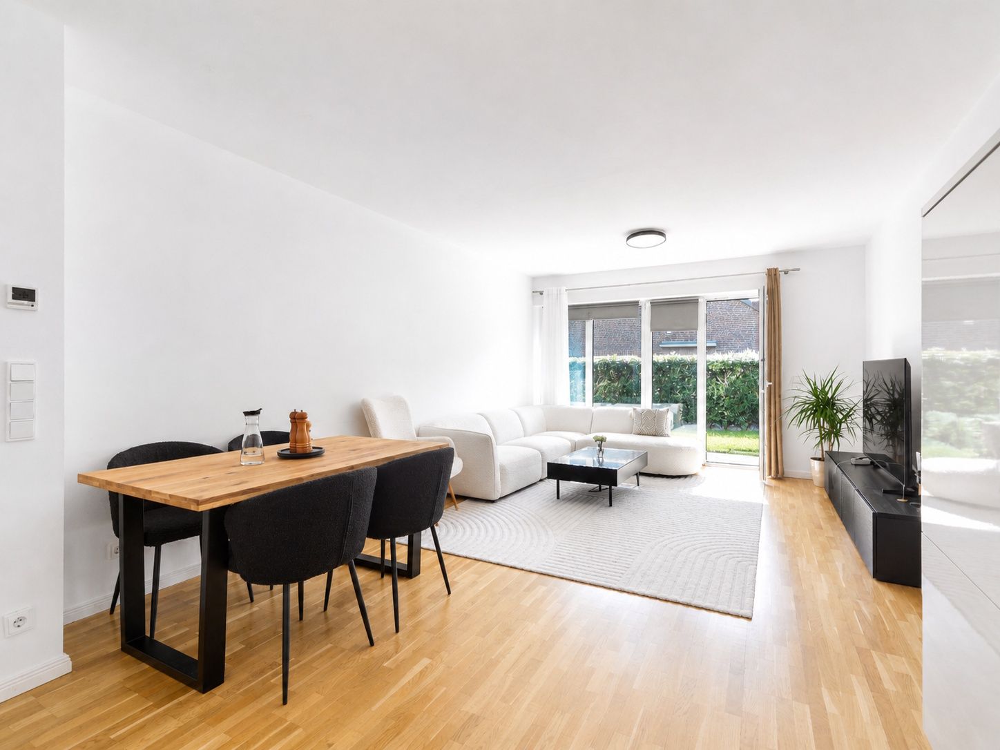 Helles Wohnzimmer mit Sofa, Esstisch, Stühlen und großem Fenster zum Garten.