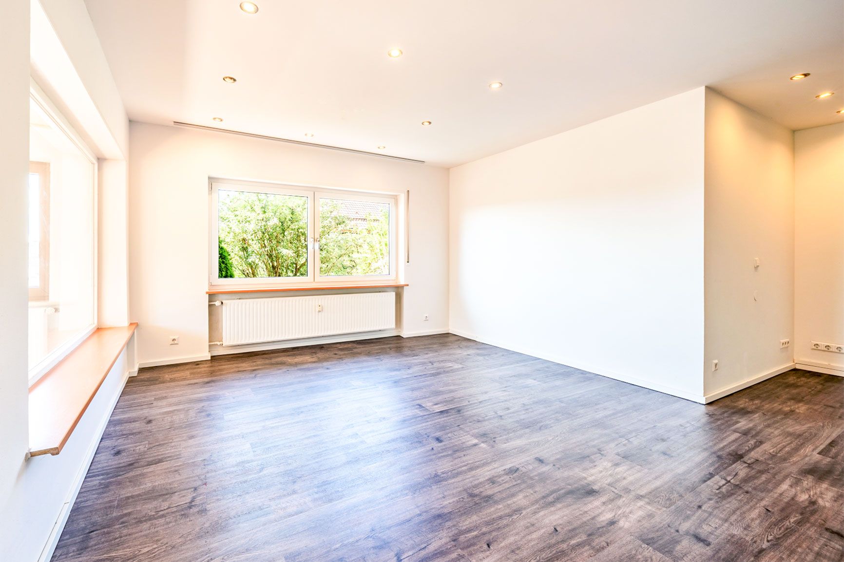 Helles, leeres Zimmer mit großem Fenster, weißer Wand und dunklem Holzboden.
