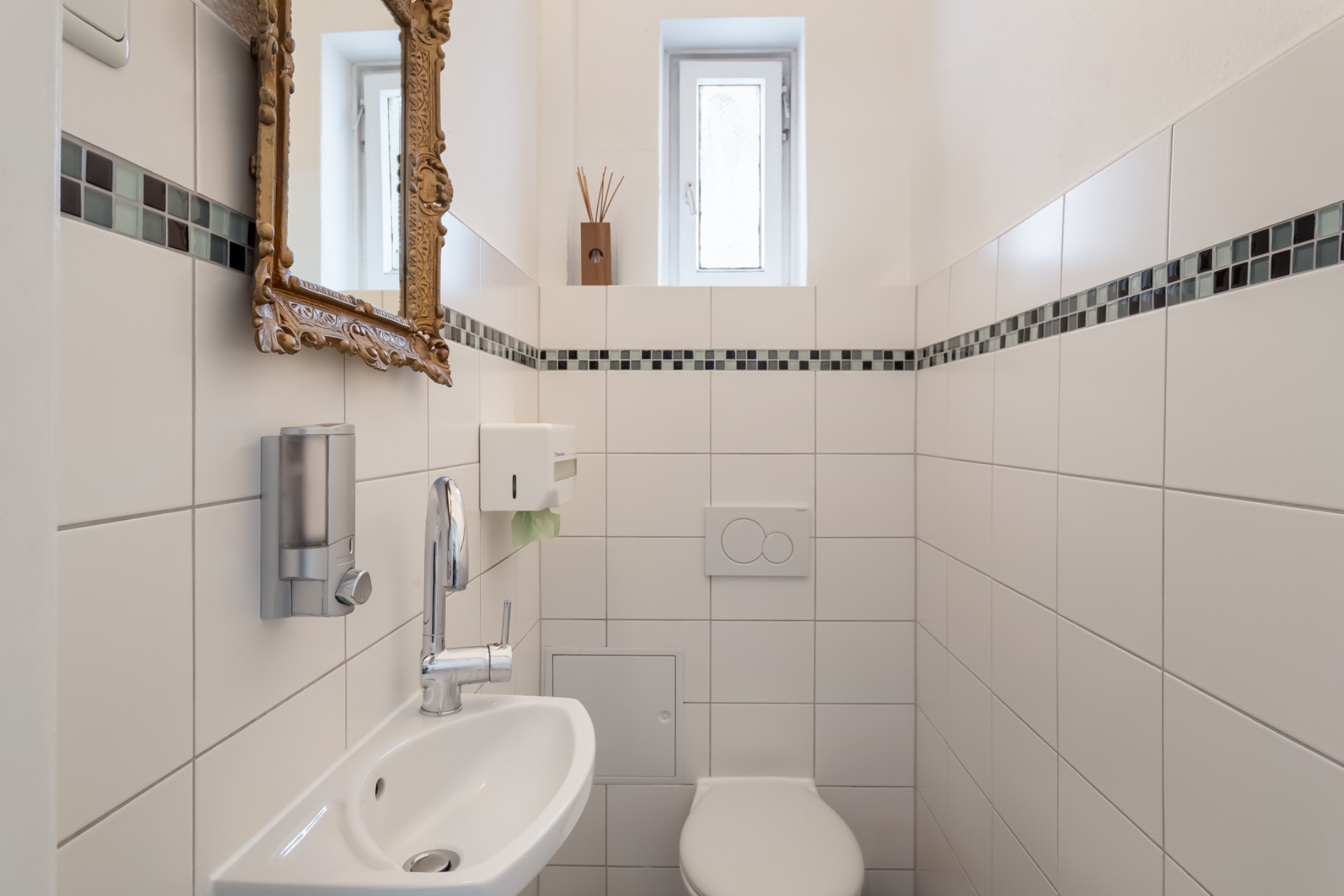 Kleines Badezimmer mit WC, Waschbecken, Spiegel und Fenster, weiße Fliesen mit Mosaikband.