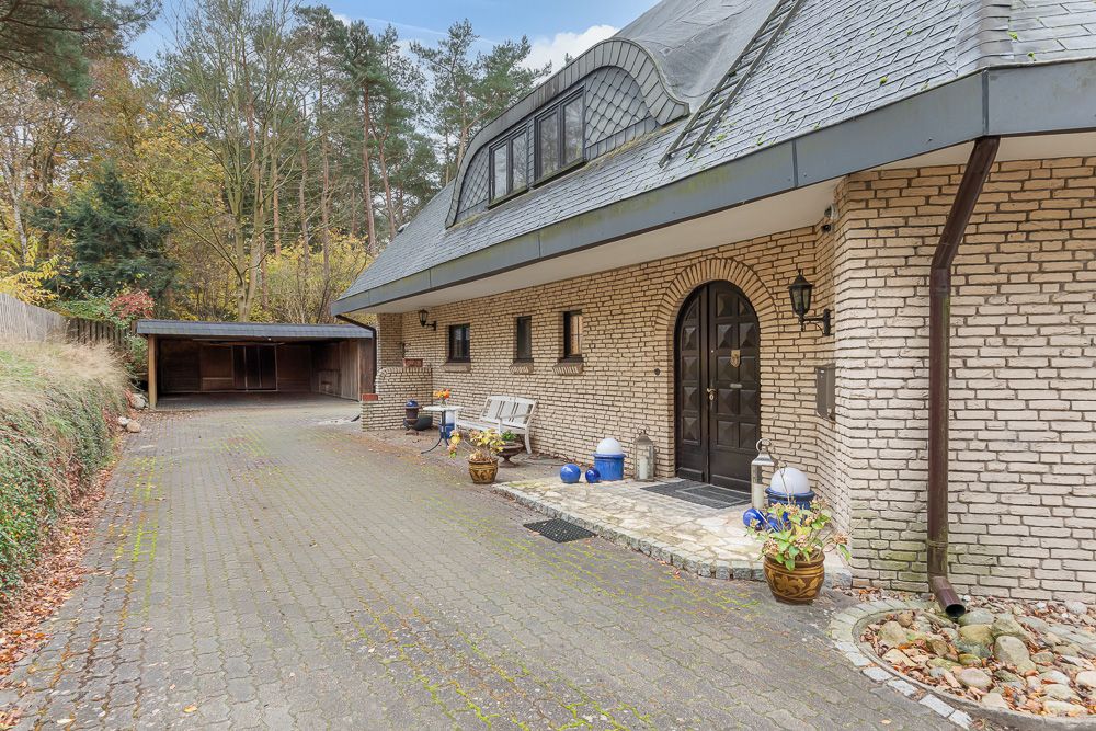 Gepflasterte Einfahrt neben einem Backsteinhaus mit Pflanzen und einer Sitzbank, Carport im Hintergrund.