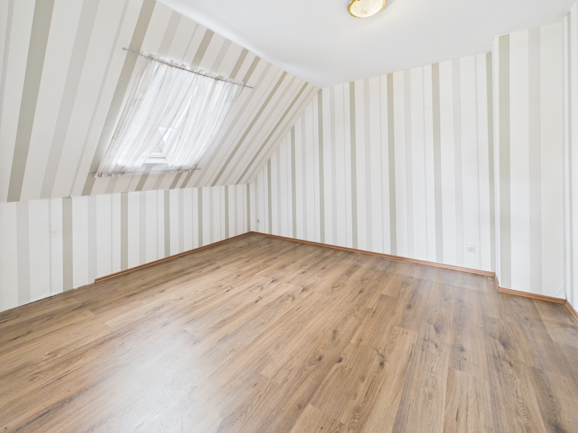 Leerer Raum mit schräger Decke, gestreiften Wänden und Fenster mit Vorhang.