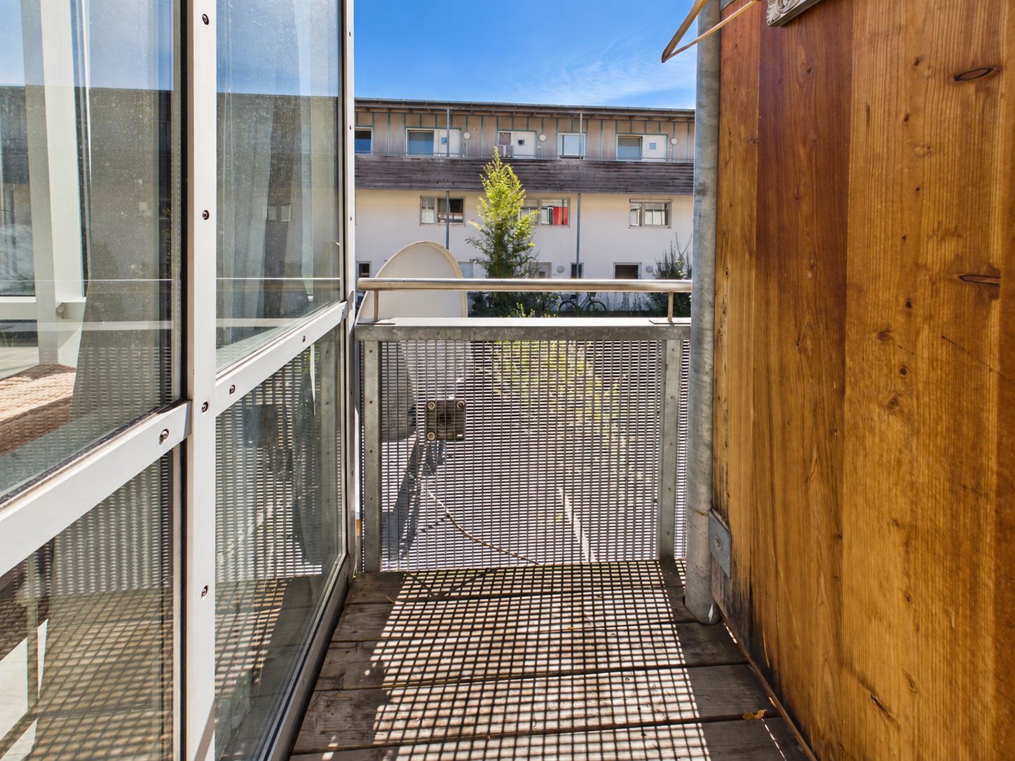 Schmaler Balkon mit Metallgeländer, Holzwand und Blick auf ein Wohnhaus bei Sonnenschein.